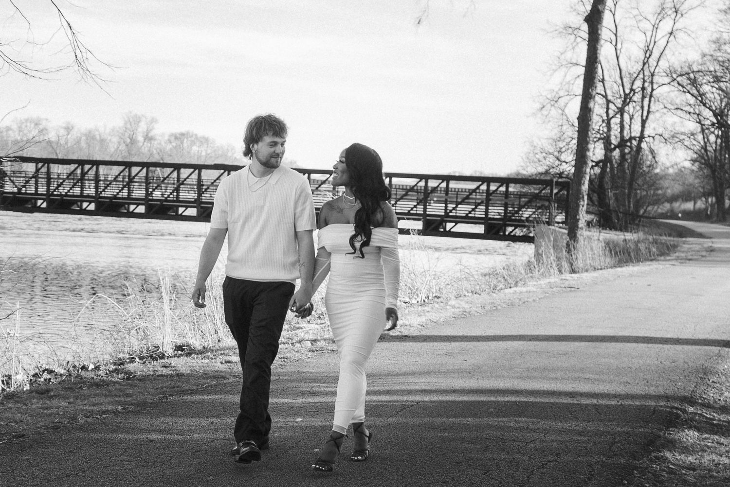 Black and white photo of a couple holding hands and walking along a path near a bridge and river.