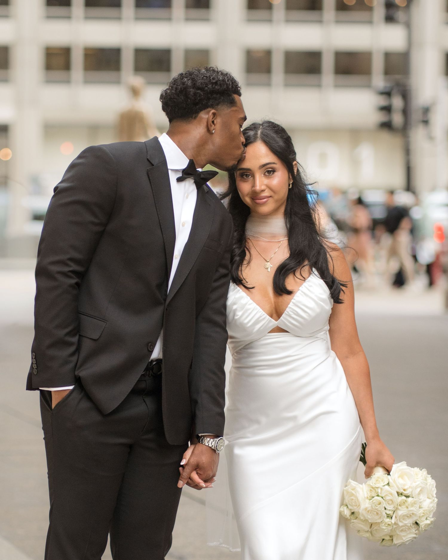 Couple in wedding attire holding hands on city street, groom in black tuxedo kissing bride in white gown holding bouquet.