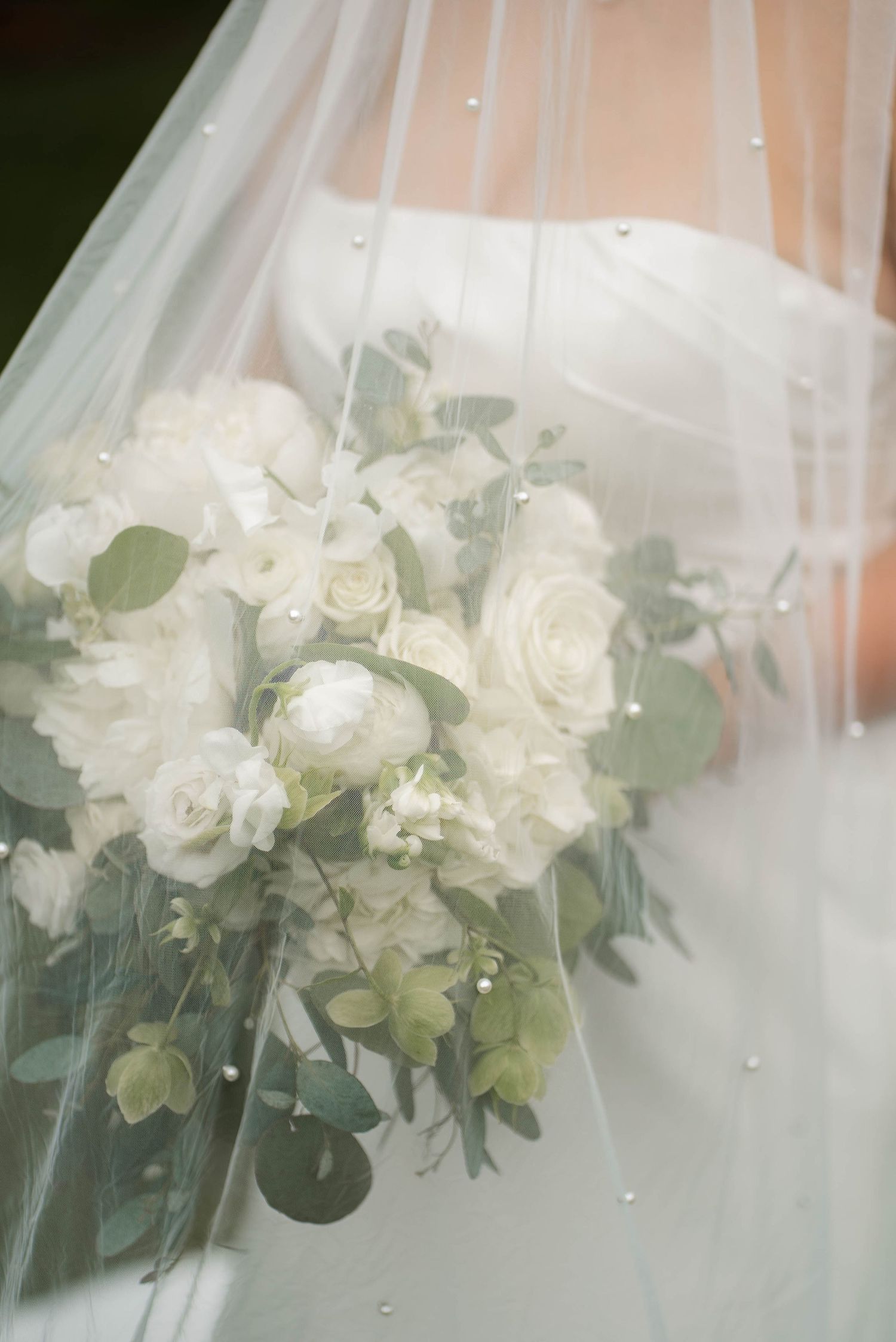 Bride holding a lush white rose and eucalyptus wedding bouquet draped with a pearl-adorned veil.