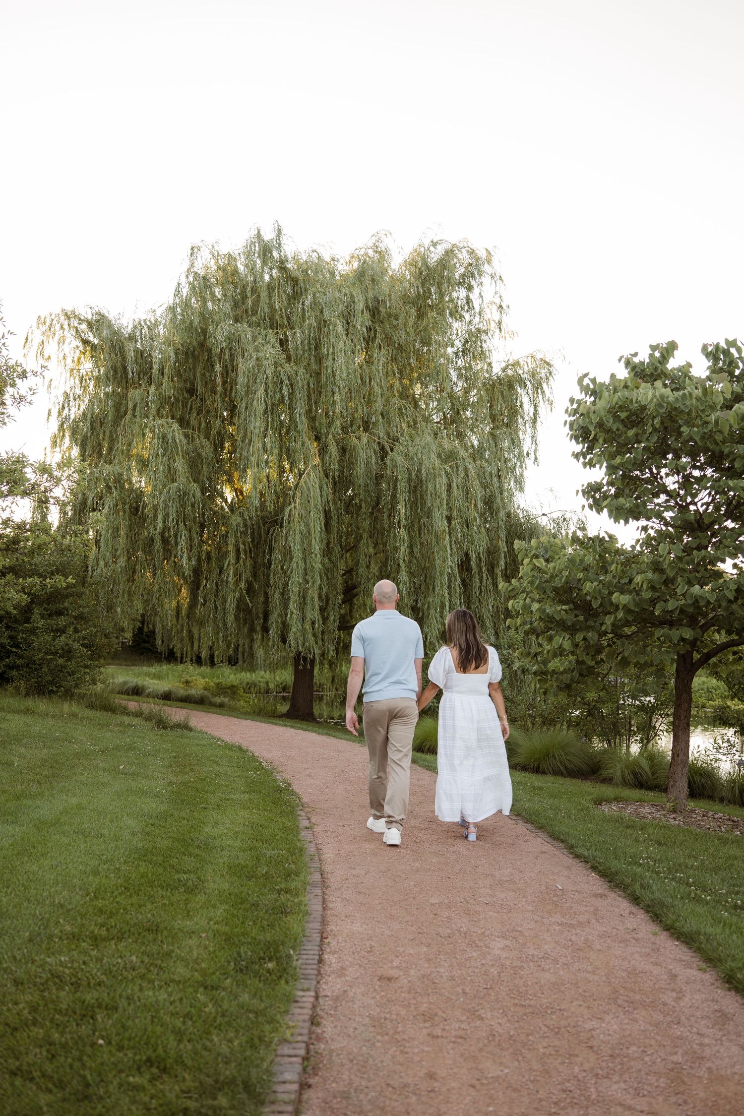 Couple walking hand in hand on a curved path toward a weeping willow tree in a lush green park.