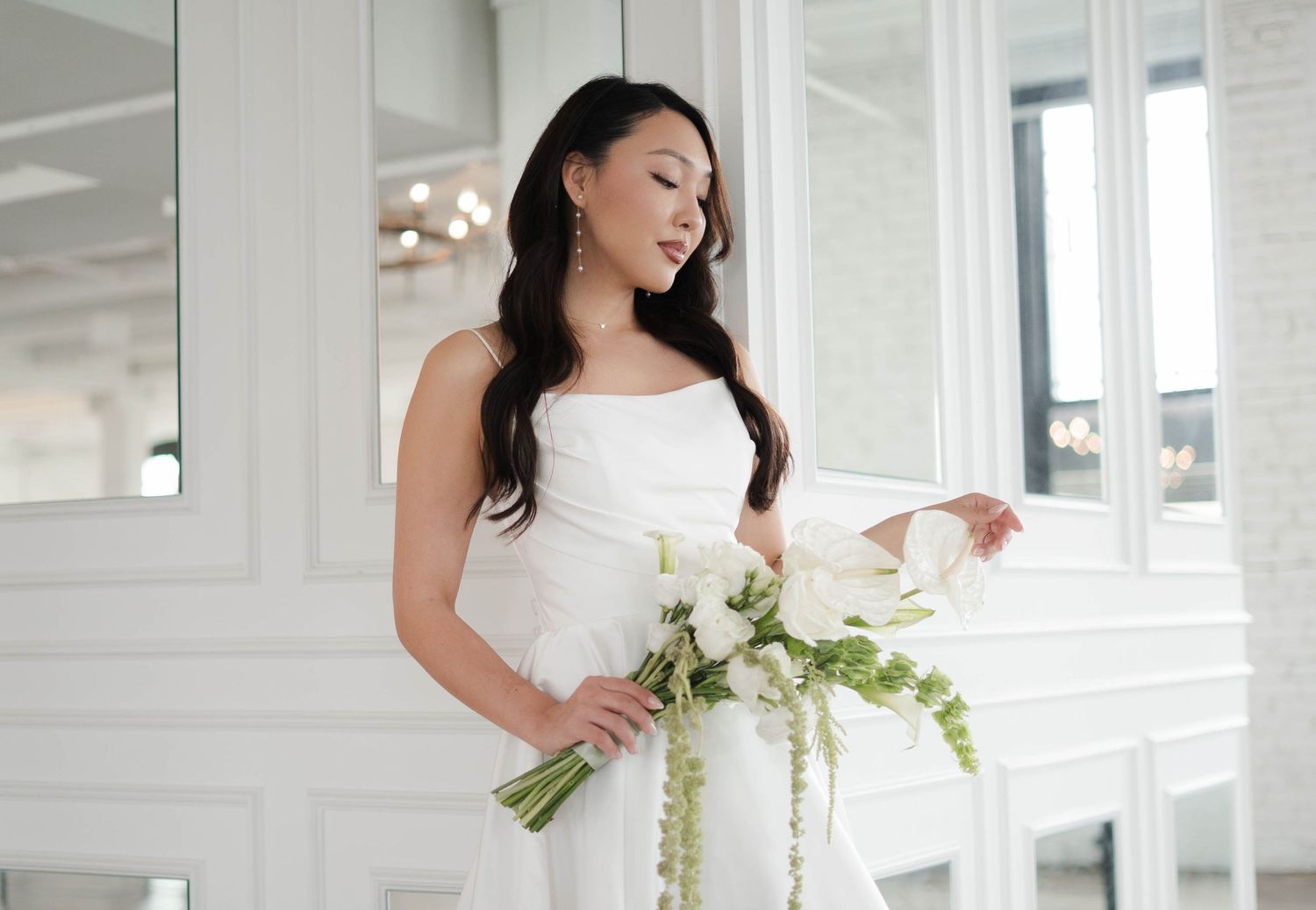 Bride in white strapless gown holding elegant white floral bouquet with greenery in a bright, modern venue.
