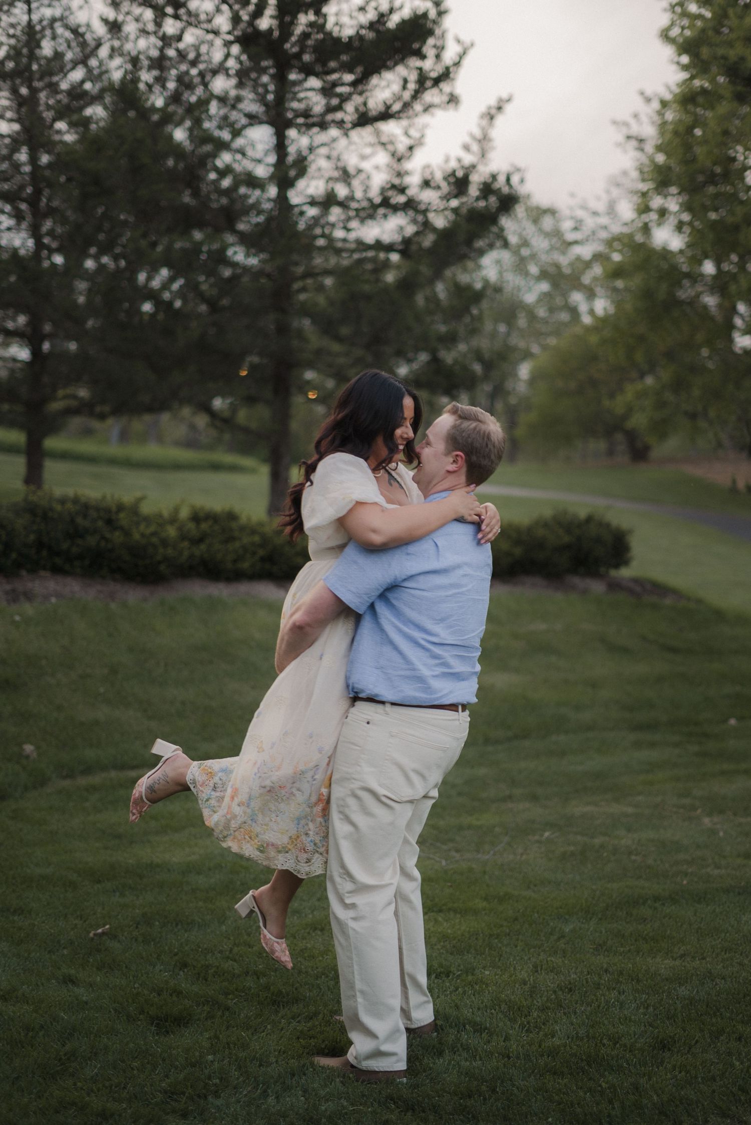 A man lifts a woman in a floral dress during a romantic outdoor engagement photo on a lush green lawn.