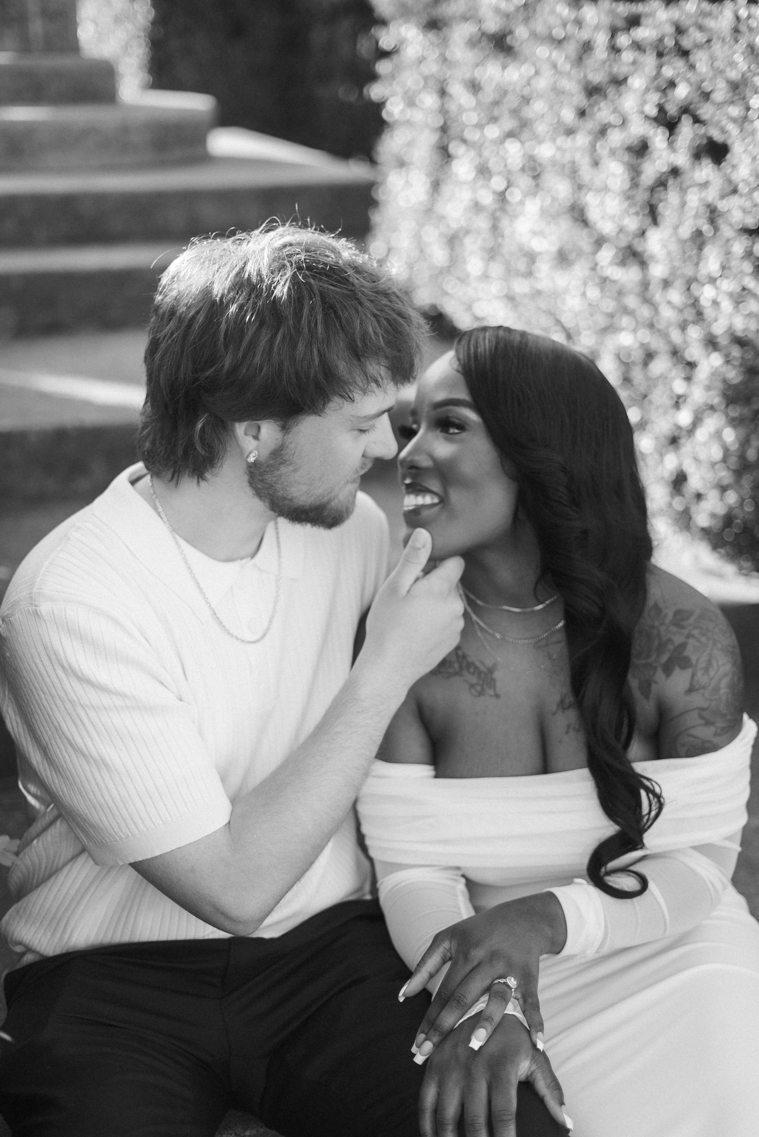 Black and white photo of a happy interracial couple sitting close together, smiling, with tattoos and elegant white outfits.