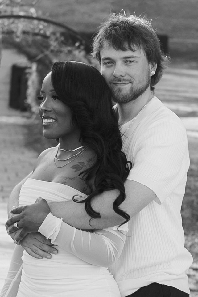 Black and white photo of a smiling couple embracing outdoors, woman in off-shoulder dress, man in white shirt.