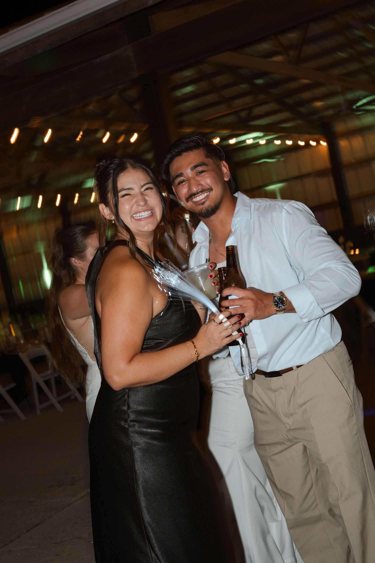 A smiling couple poses at an evening wedding reception, woman in black dress holding champagne bottle, man in white shirt holding beer.