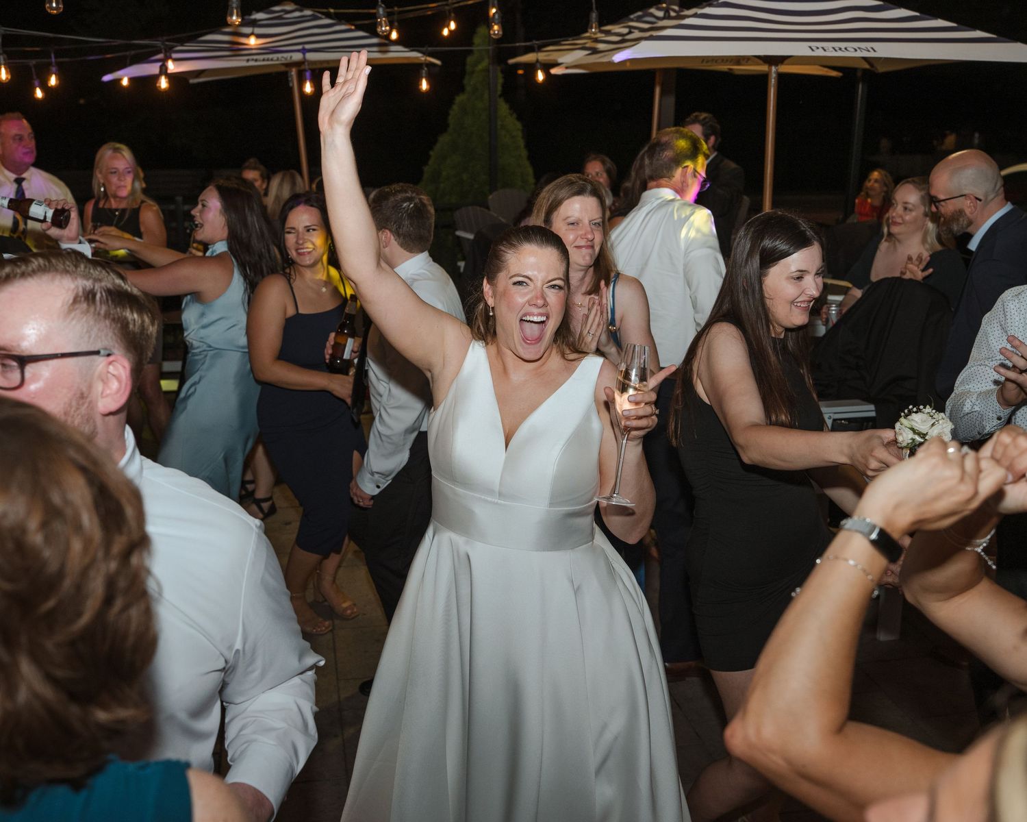 Joyful bride in white gown dancing with raised arms at outdoor wedding reception surrounded by guests.