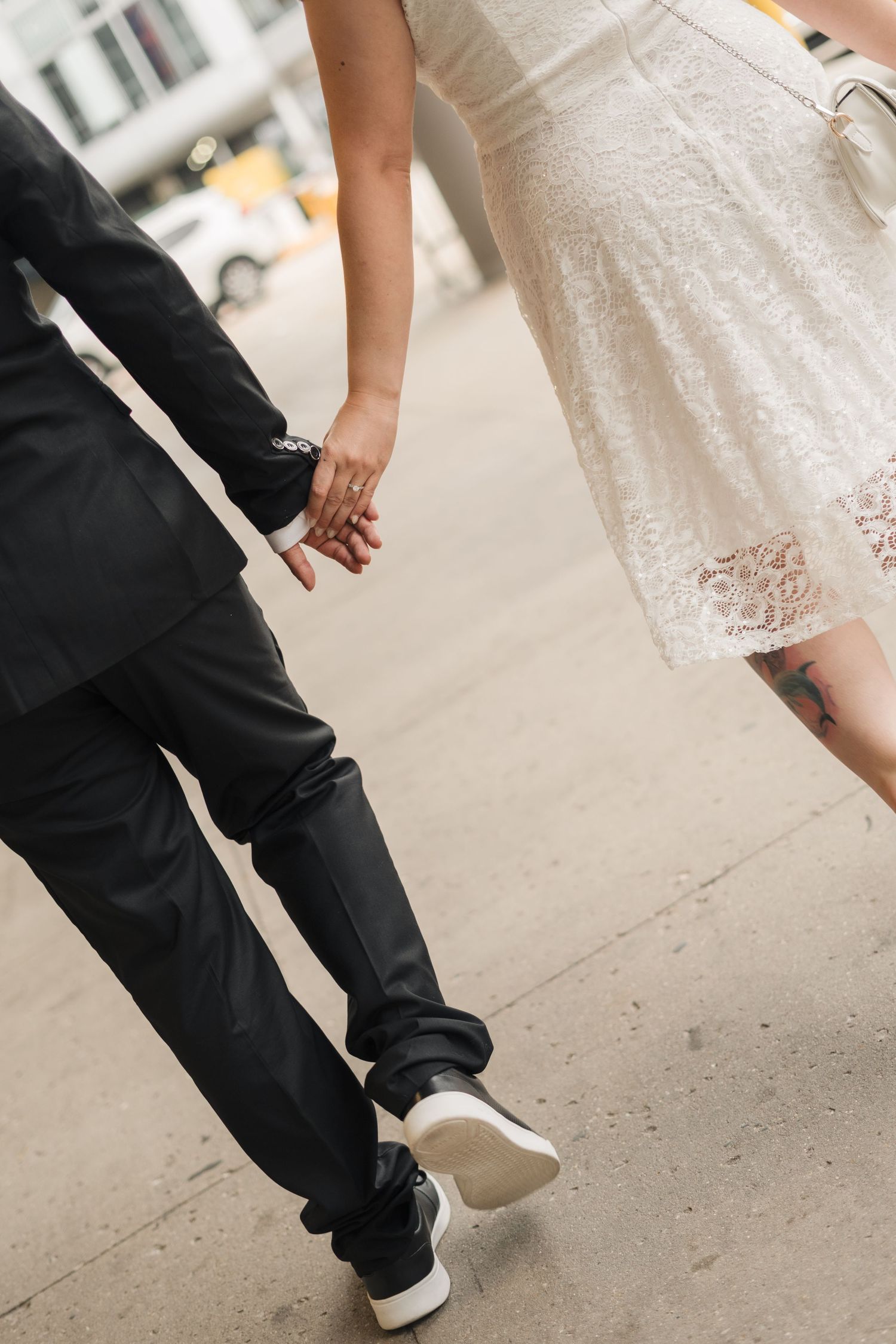 Couple holding hands walking on sidewalk, groom in black suit and sneakers, bride in white lace dress with tattoo.