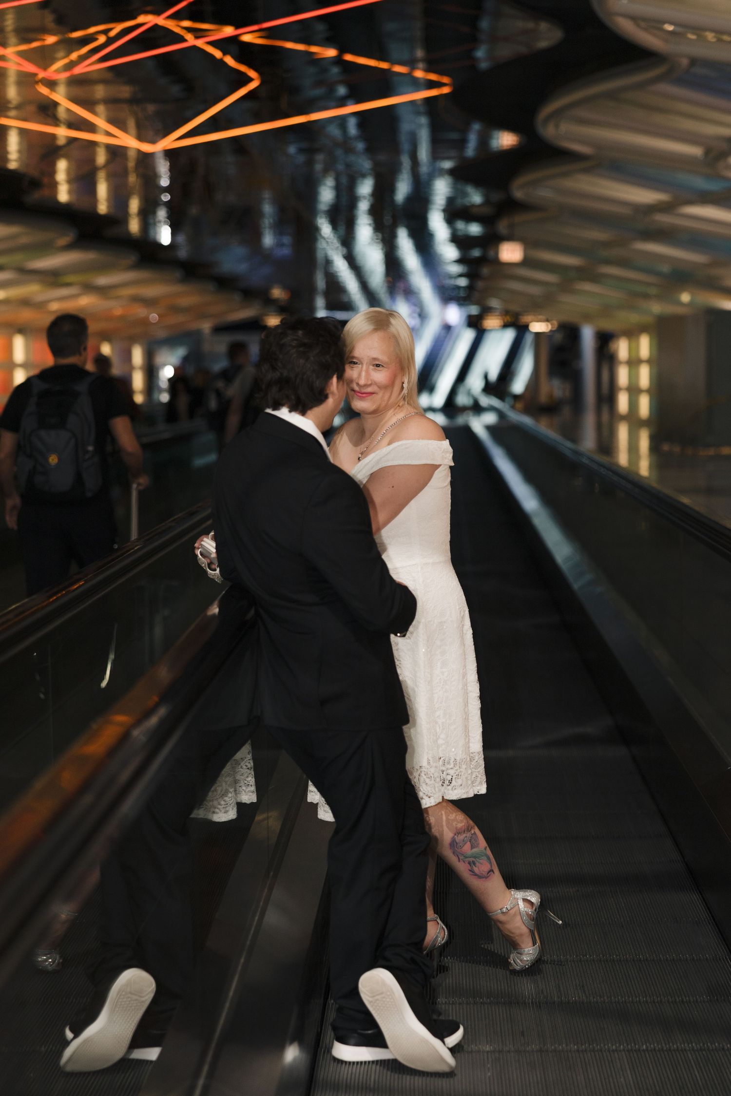 Couple in wedding attire embracing on an escalator in a modern venue, woman in white dress with tattoo, man in black suit.