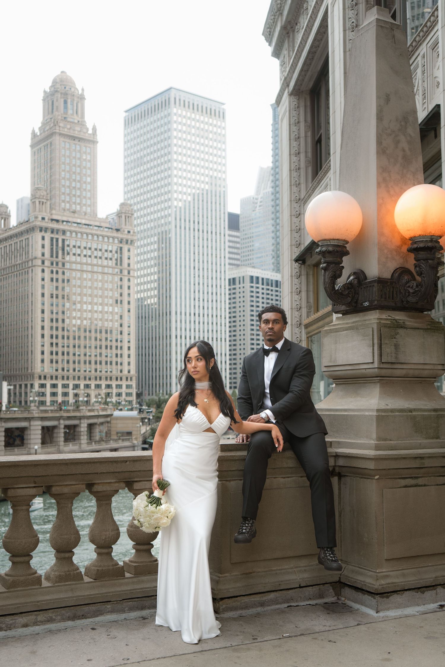 Wedding couple posing on a bridge with Chicago skyline backdrop, bride in white gown and groom in black tuxedo.
