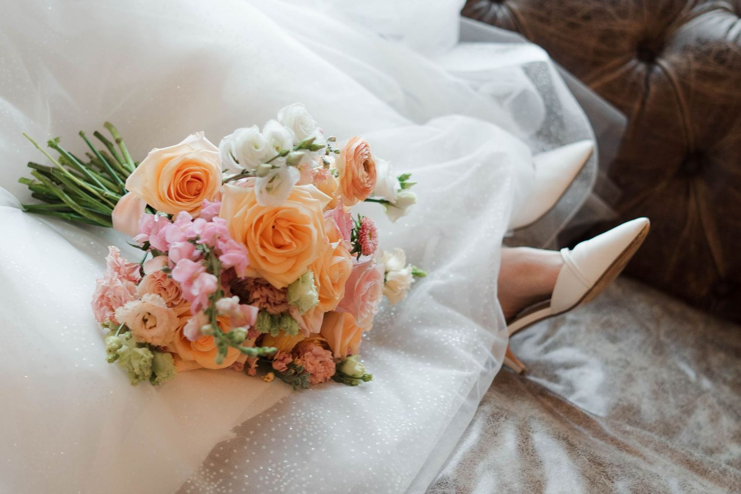 Bridal bouquet of peach roses and pink flowers beside white heels on a wedding dress.