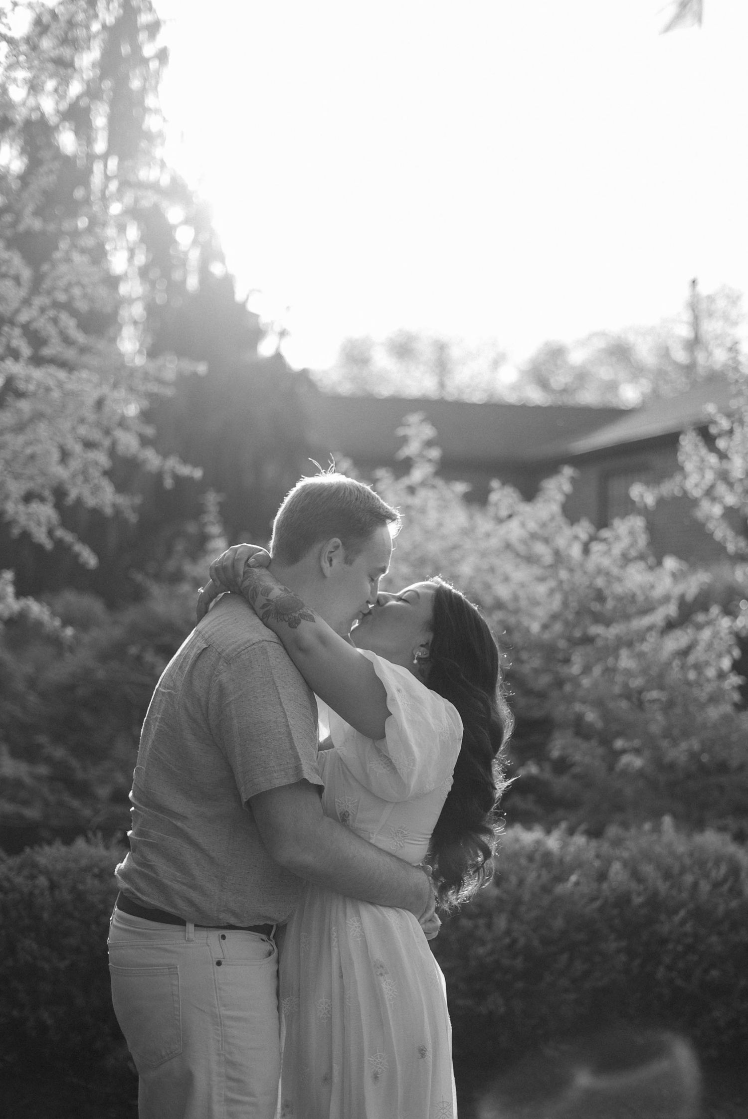 Black and white photo of a couple embracing and about to kiss in a lush garden setting with soft backlight.