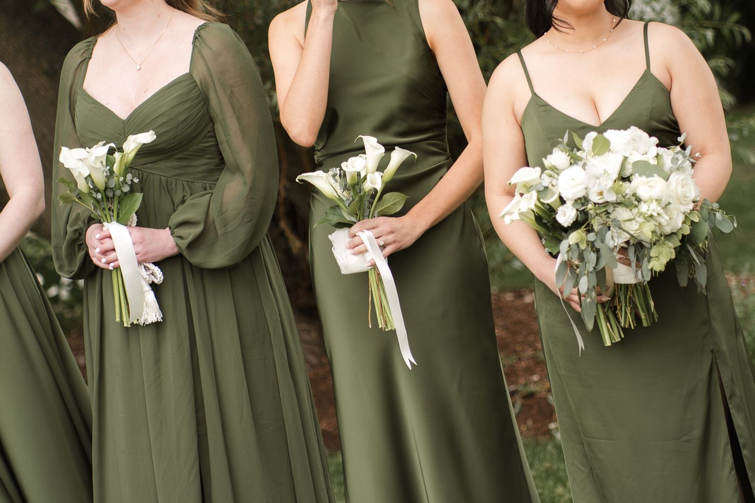 Bridesmaids in olive green gowns holding white floral bouquets with ribbon ties at an outdoor wedding.