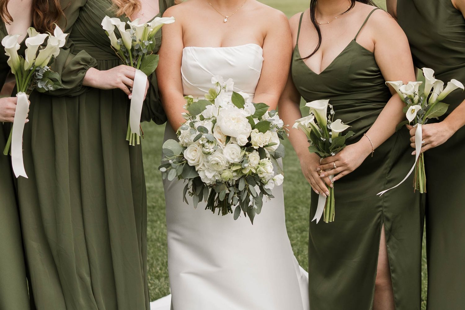 Bride in white gown holding white floral bouquet flanked by bridesmaids in olive green dresses with matching bouquets.