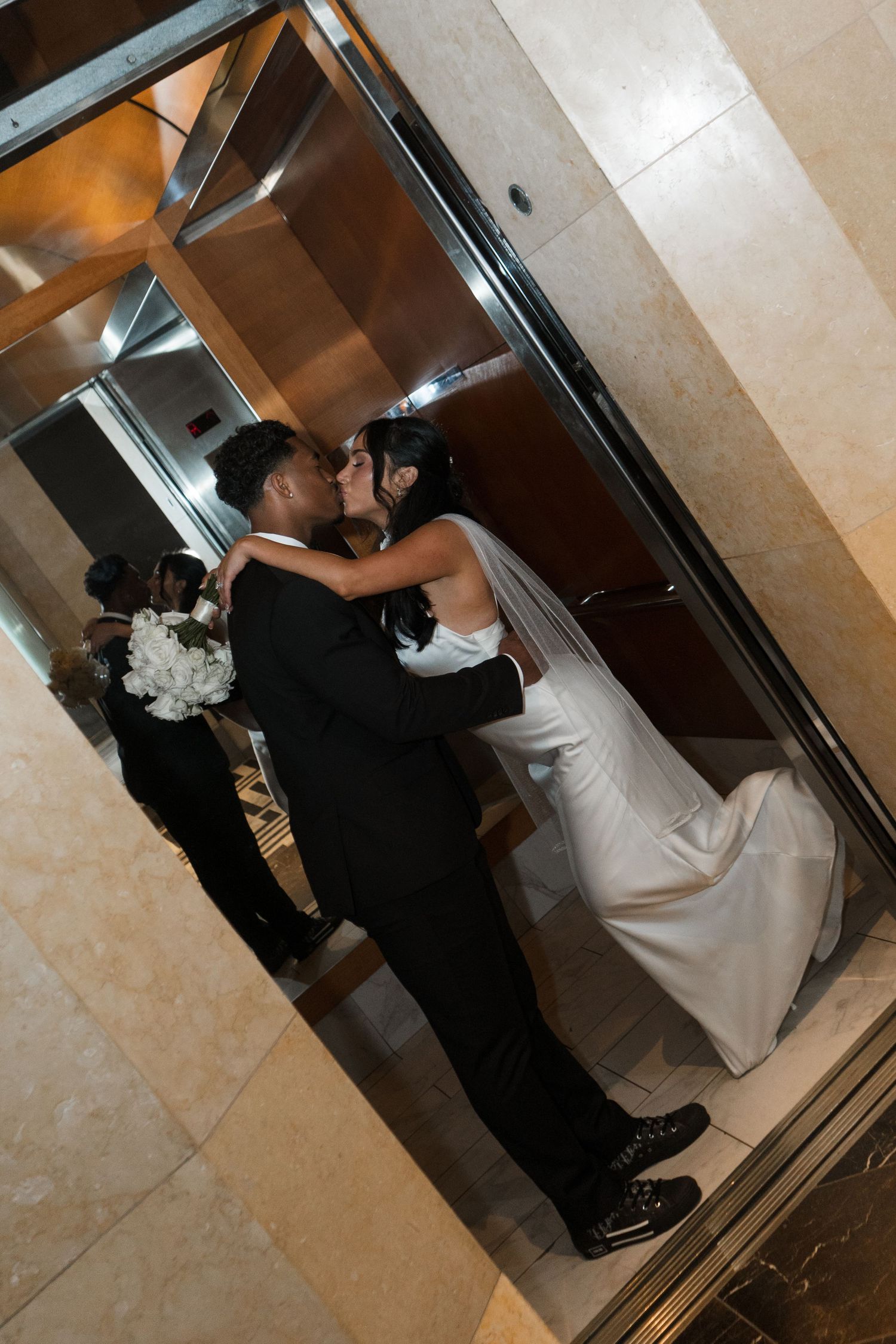 Newlywed couple sharing a kiss in an elevator, bride in white gown with veil, groom in black tuxedo.