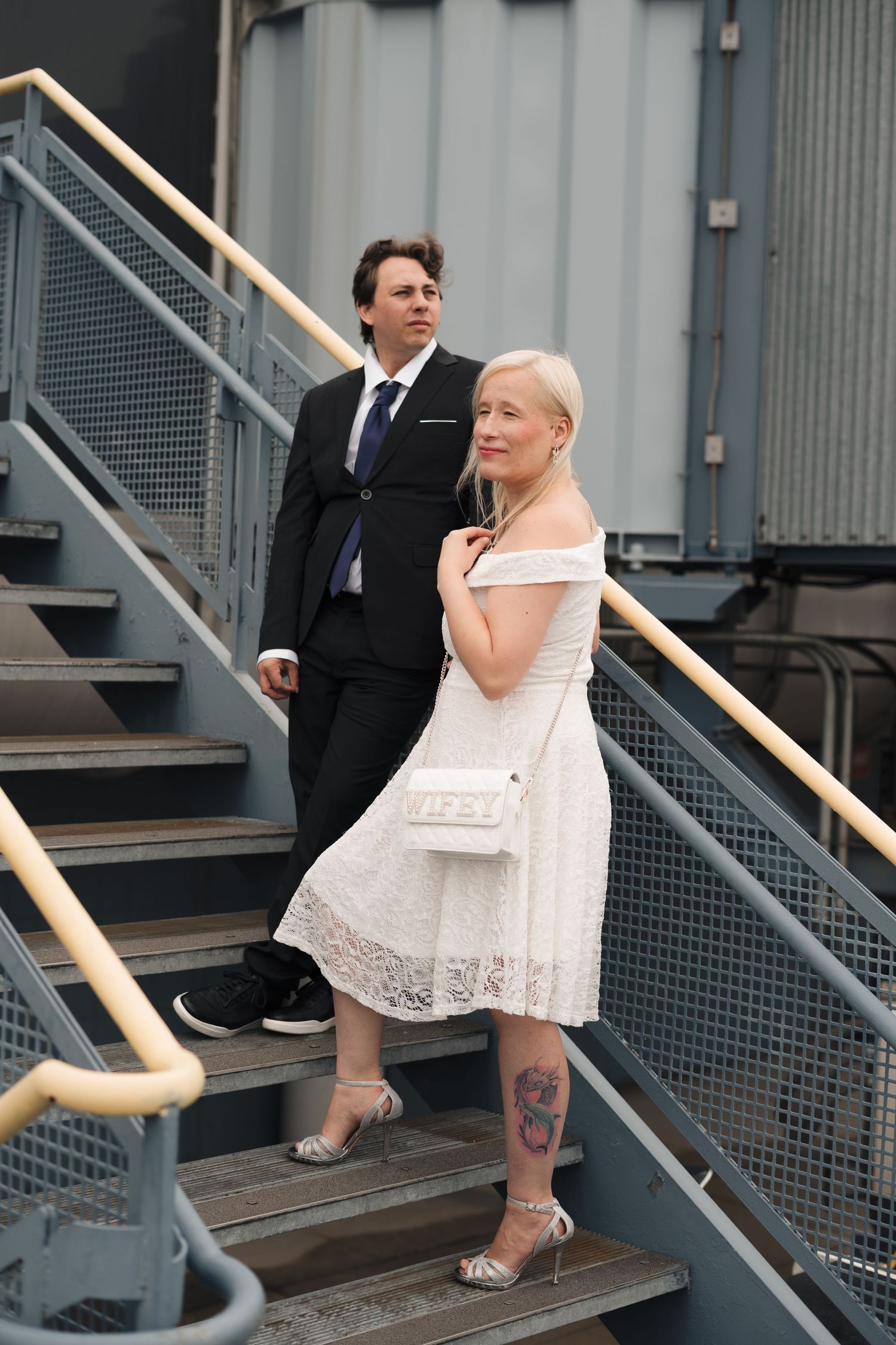 Couple in wedding attire on industrial stairs, woman in white lace dress with tattoo, man in black suit behind her.