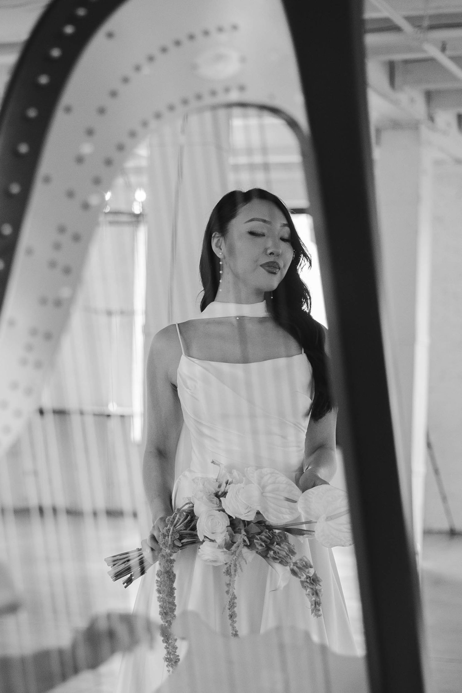 Black and white photo of a bride in a white dress holding a floral bouquet, framed through harp strings indoors.