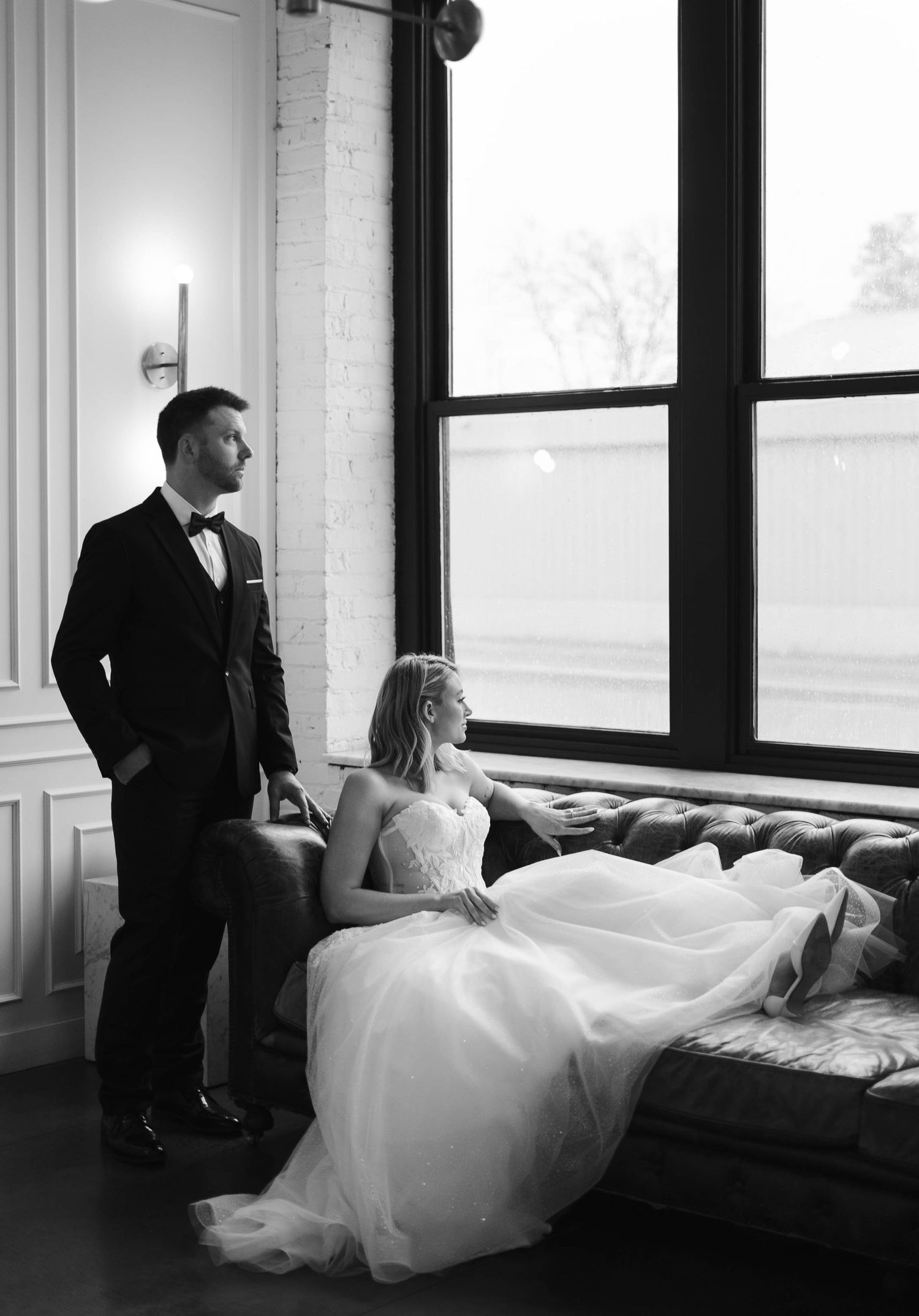 Black and white photo of a bride in a flowing gown seated on a couch beside a groom in a tuxedo near large windows.