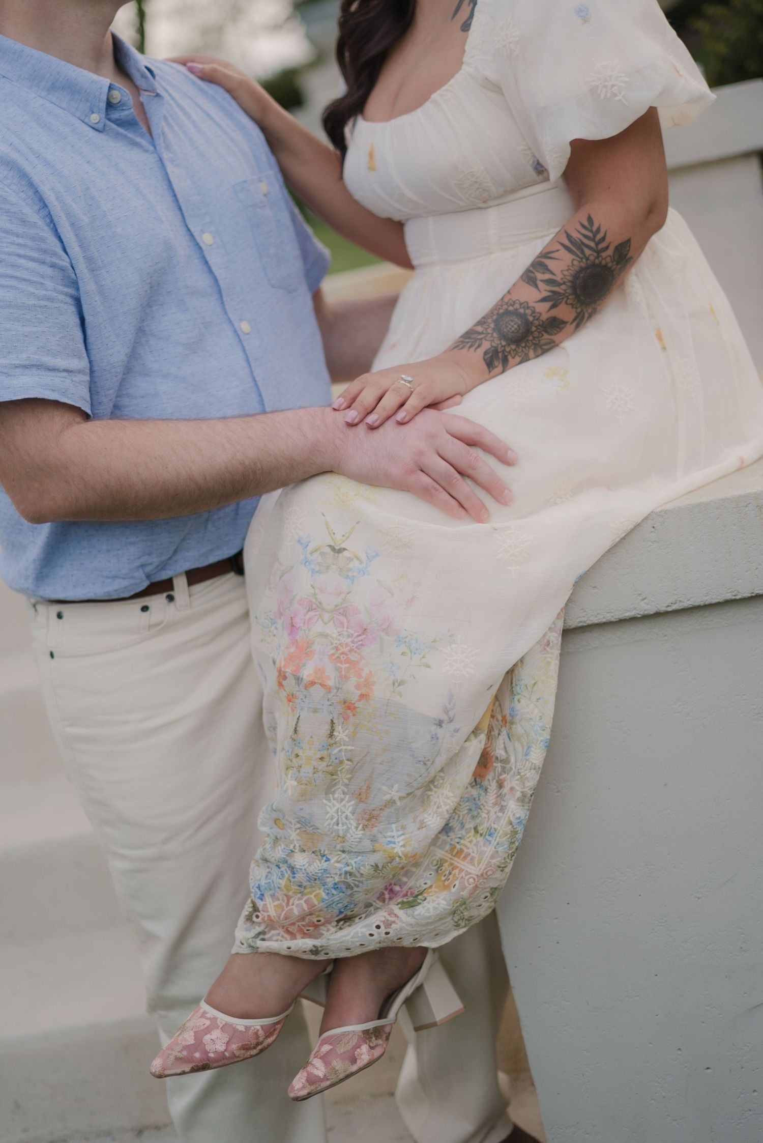Couple posing together, woman in floral dress with tattoos, man in blue shirt, hands gently touching.