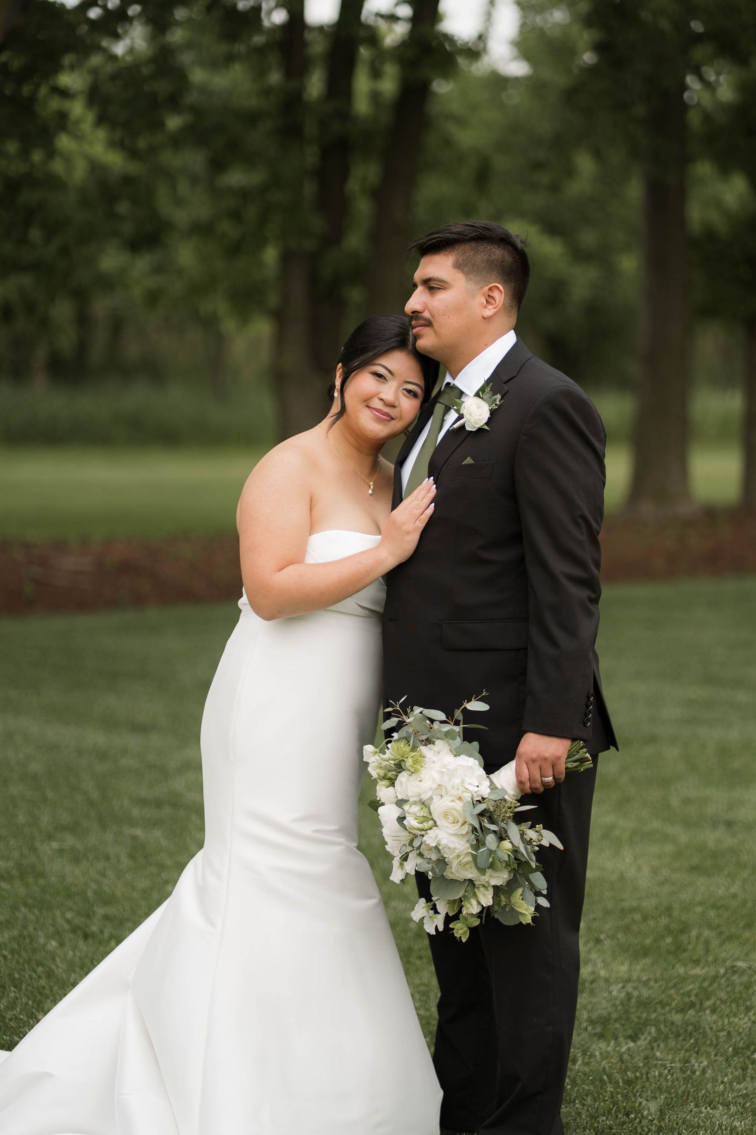 Bride in strapless mermaid gown embraces groom in black tuxedo outdoors holding white floral bouquet.