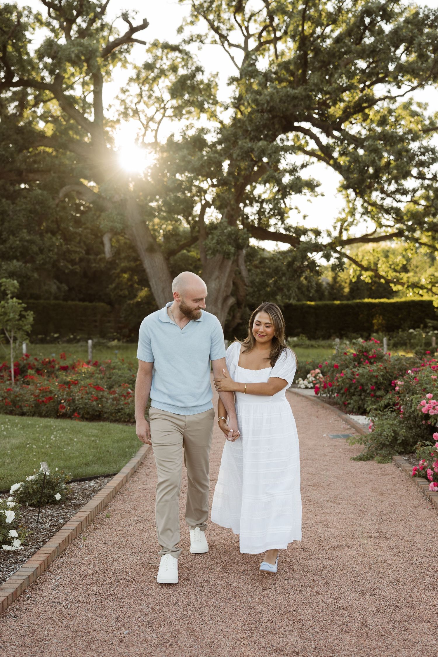 Couple walking hand in hand through rose garden path at golden hour during outdoor engagement session.