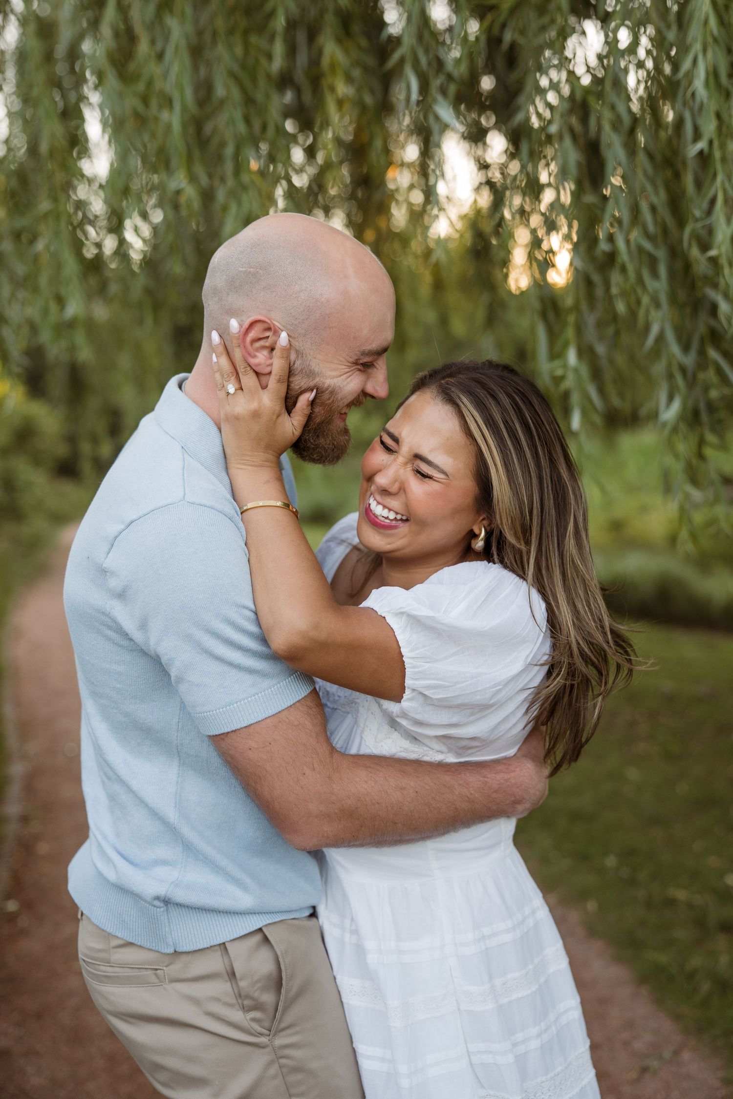 Happy couple embracing and laughing under a weeping willow tree during a romantic outdoor engagement session.