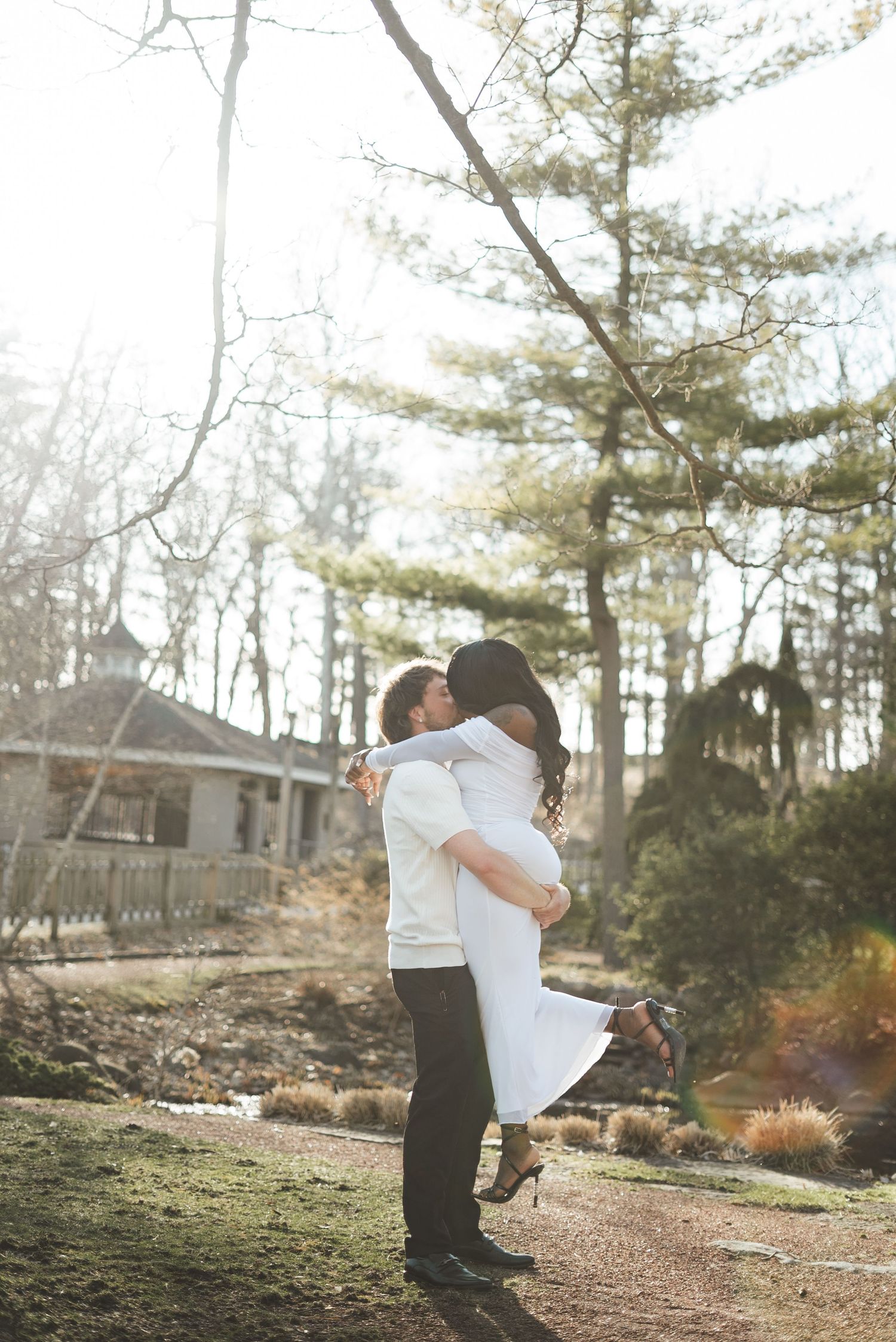 A man lifts a woman in a white dress for a romantic kiss outdoors in a sunlit garden with trees in the background.