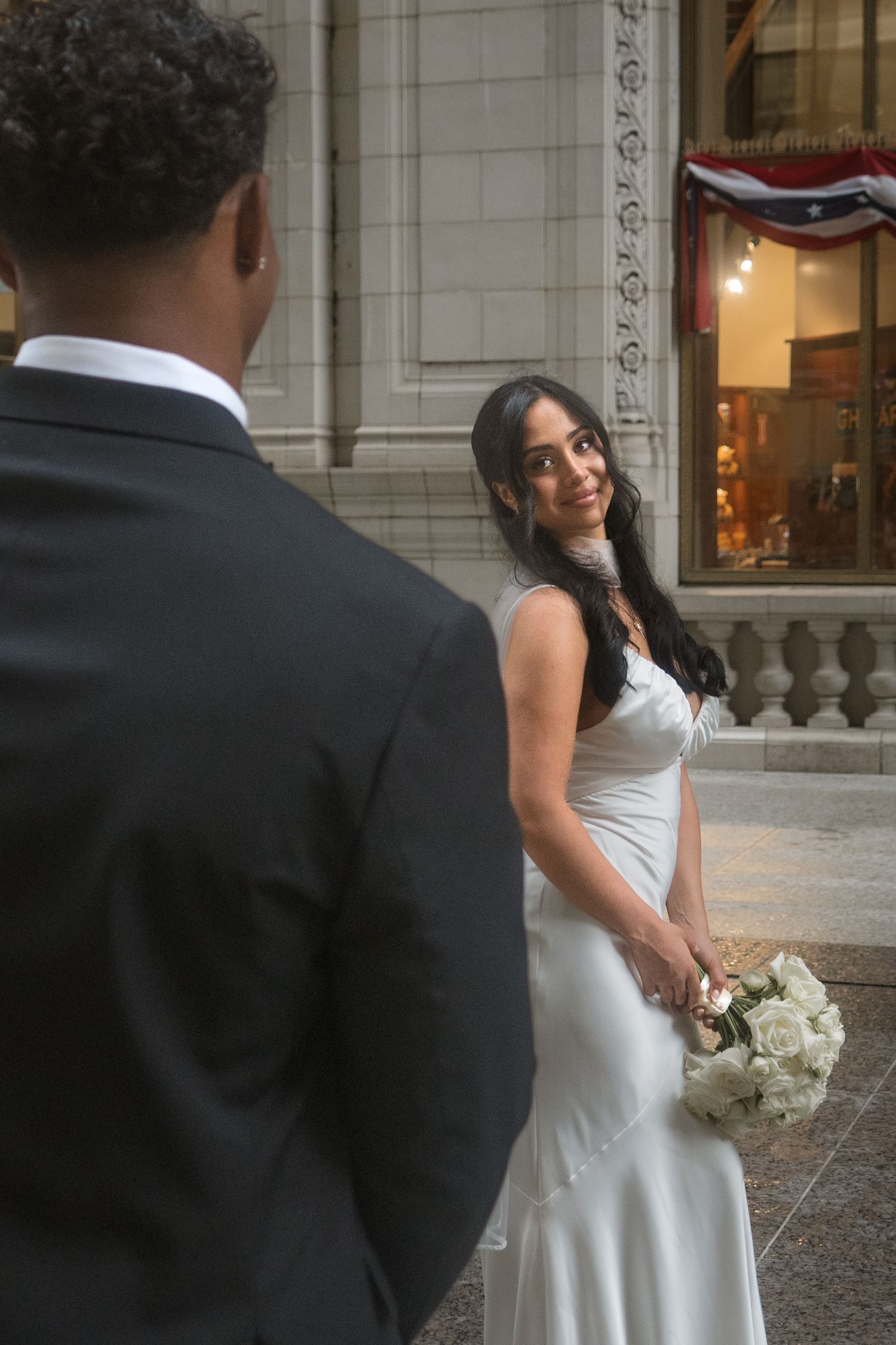 Bride in white gown holds bouquet, smiling at groom in black suit on elegant building steps.