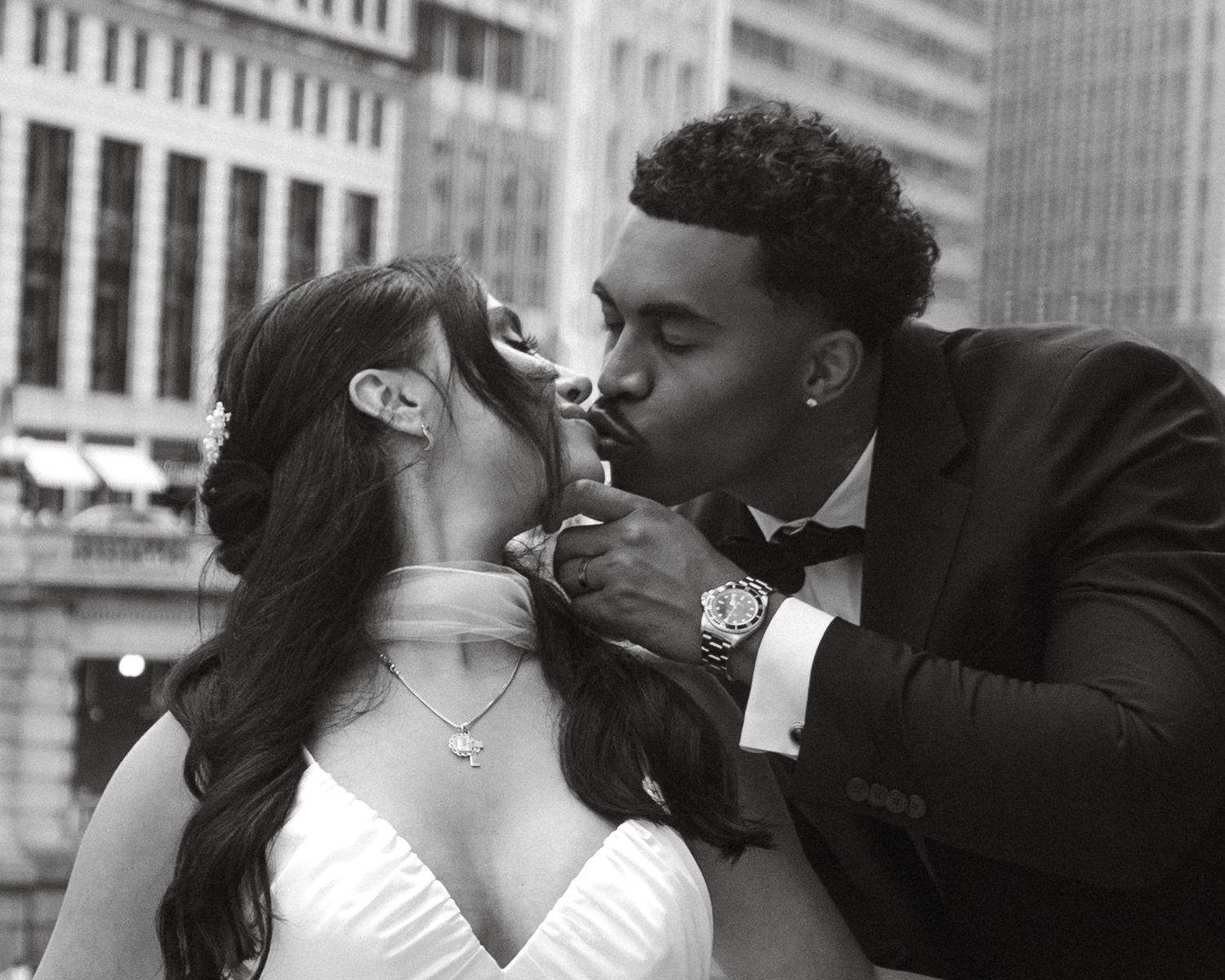 Black and white photo of a couple sharing a kiss, bride in white gown and groom in tuxedo with city backdrop.