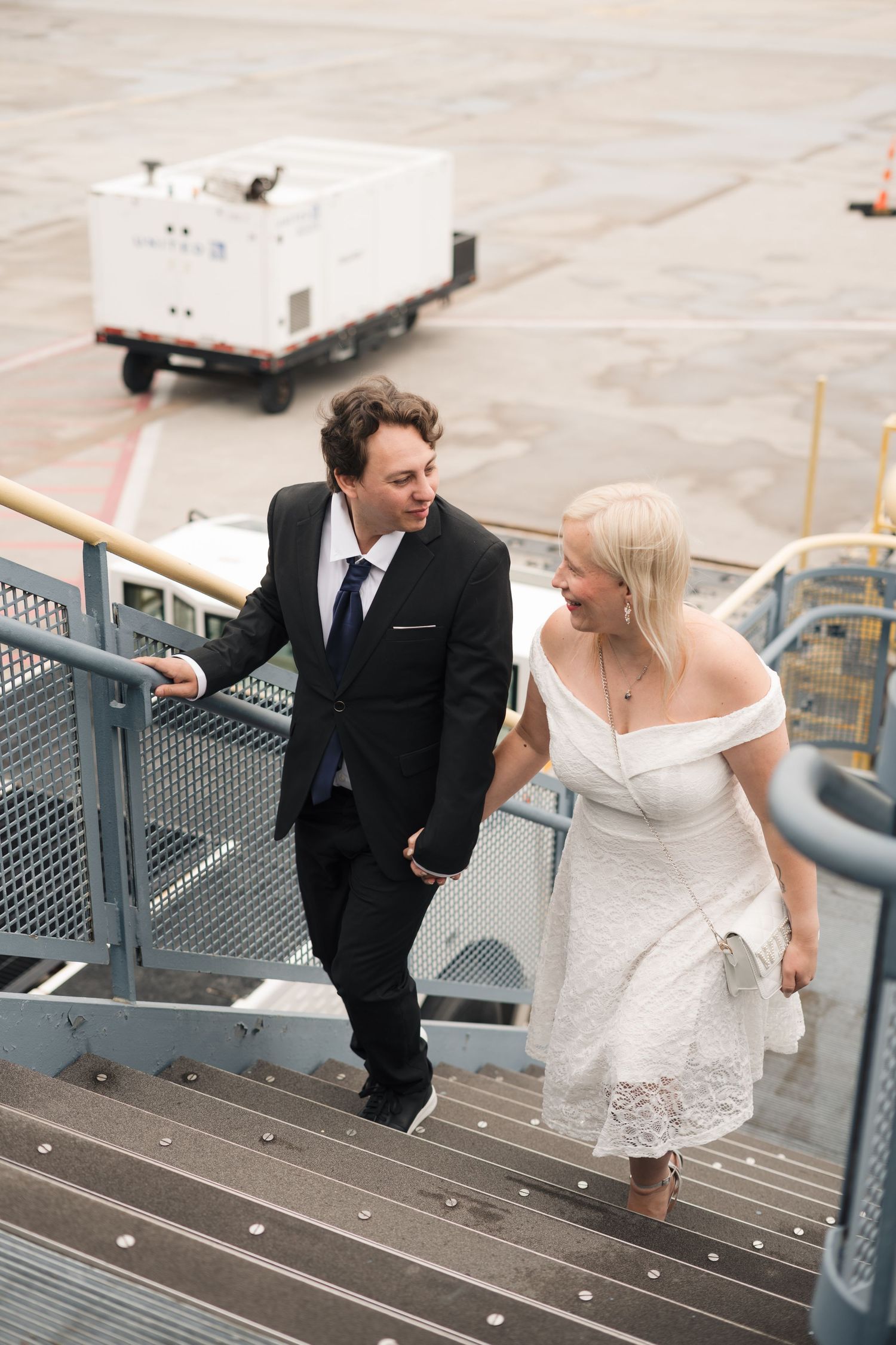 Newlywed couple in wedding attire holding hands while climbing airport stairs on tarmac.