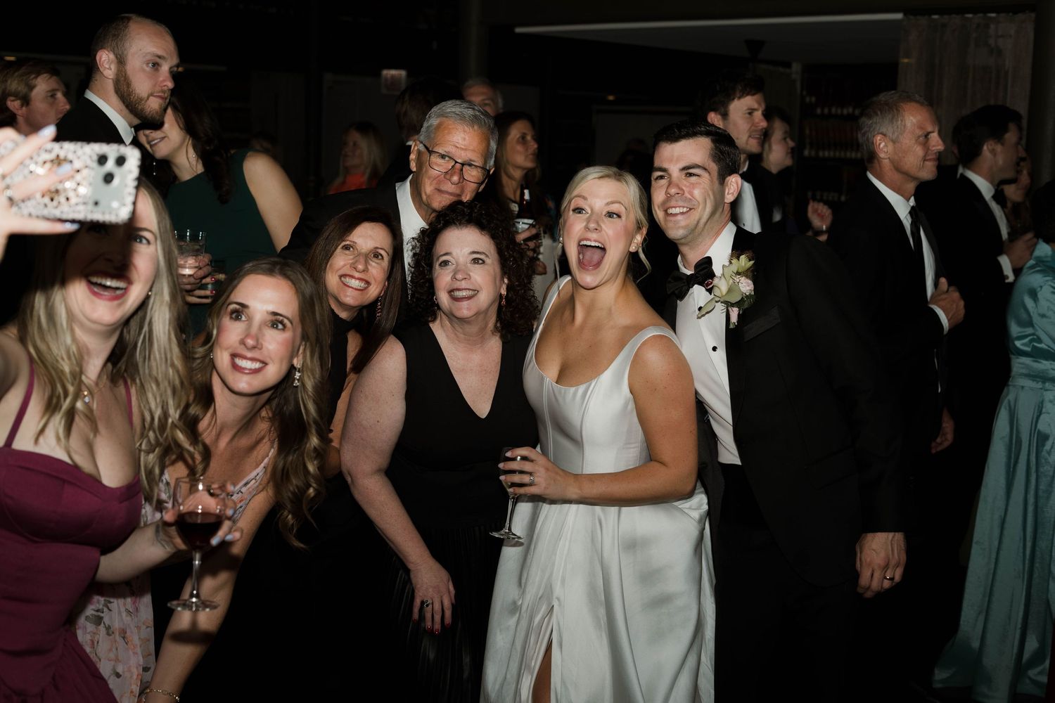 Bride in white gown poses with smiling wedding guests on dance floor at elegant evening reception.