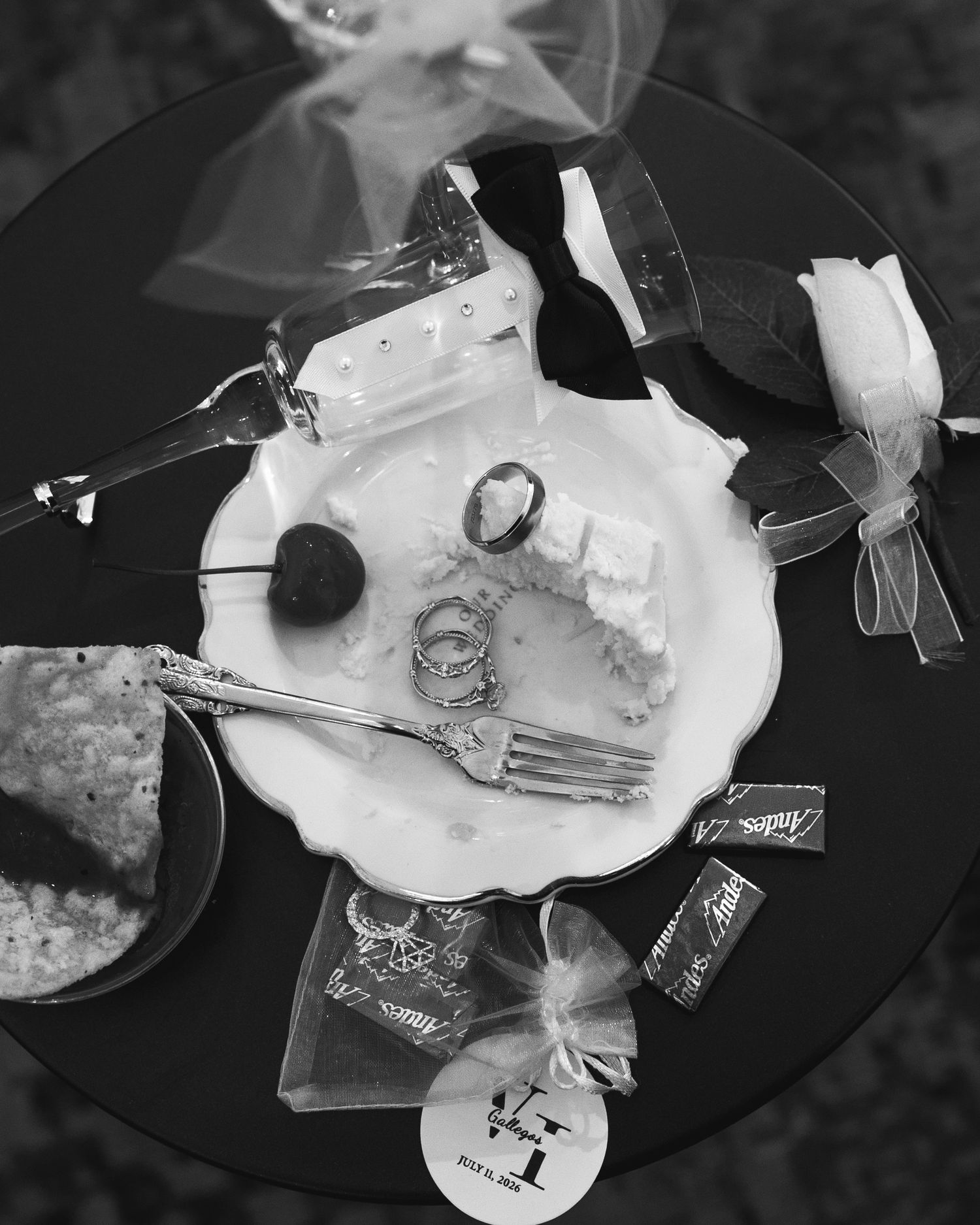 Black and white photo of a wedding cake slice on decorative plate with fork, rings, and gift favors on round table.