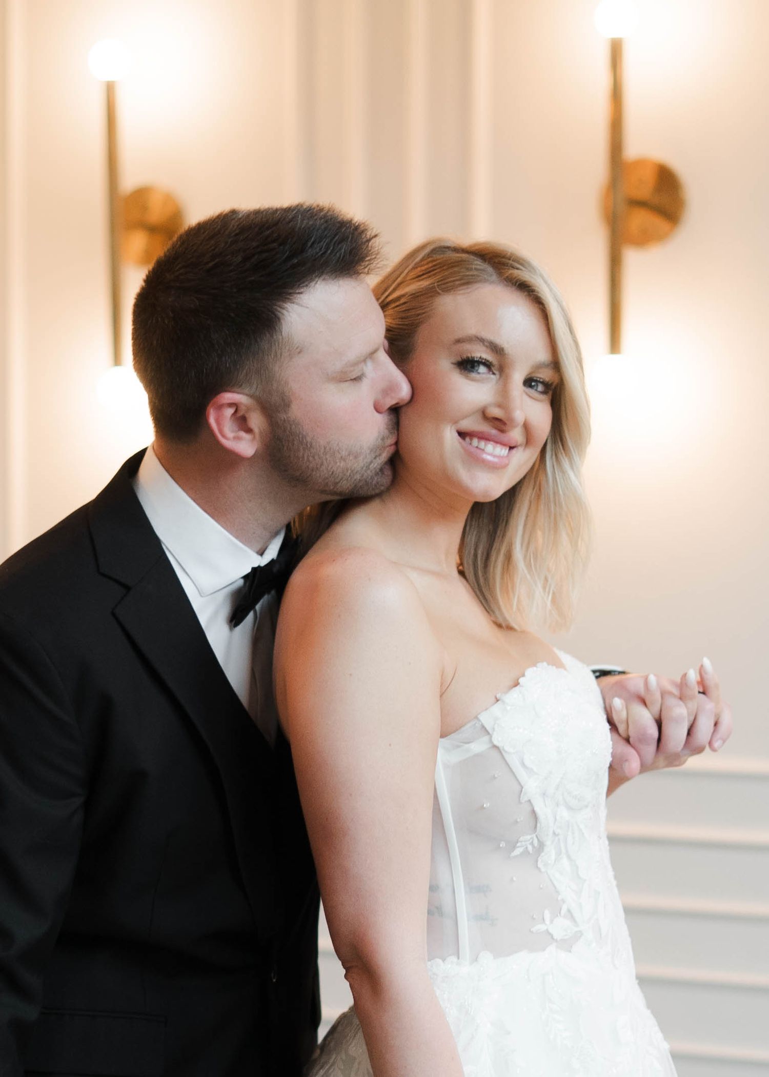 A groom in a black tuxedo kisses his smiling bride in a white strapless gown.
