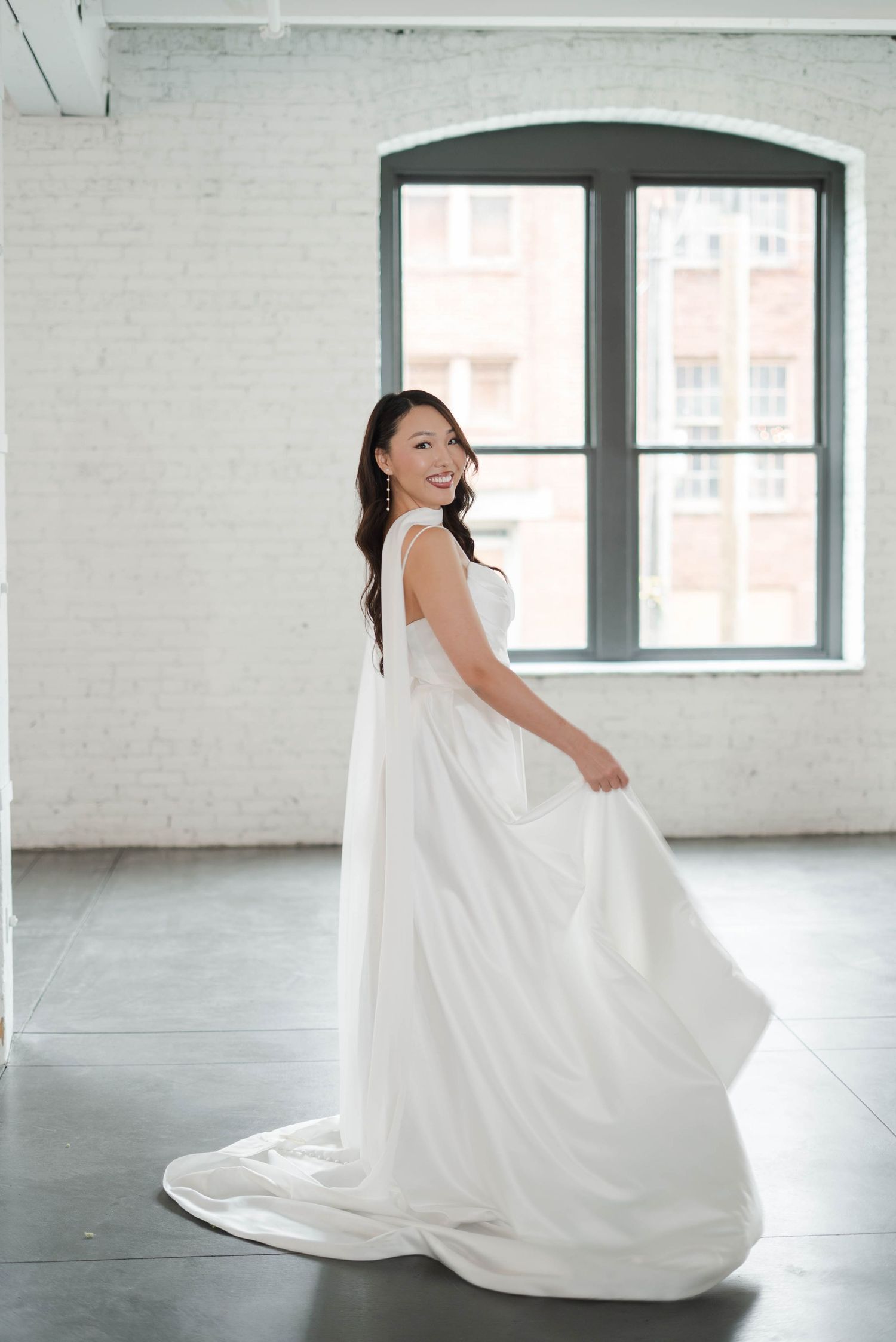 Smiling bride in elegant white ball gown twirling in a bright studio with white brick walls and large windows.
