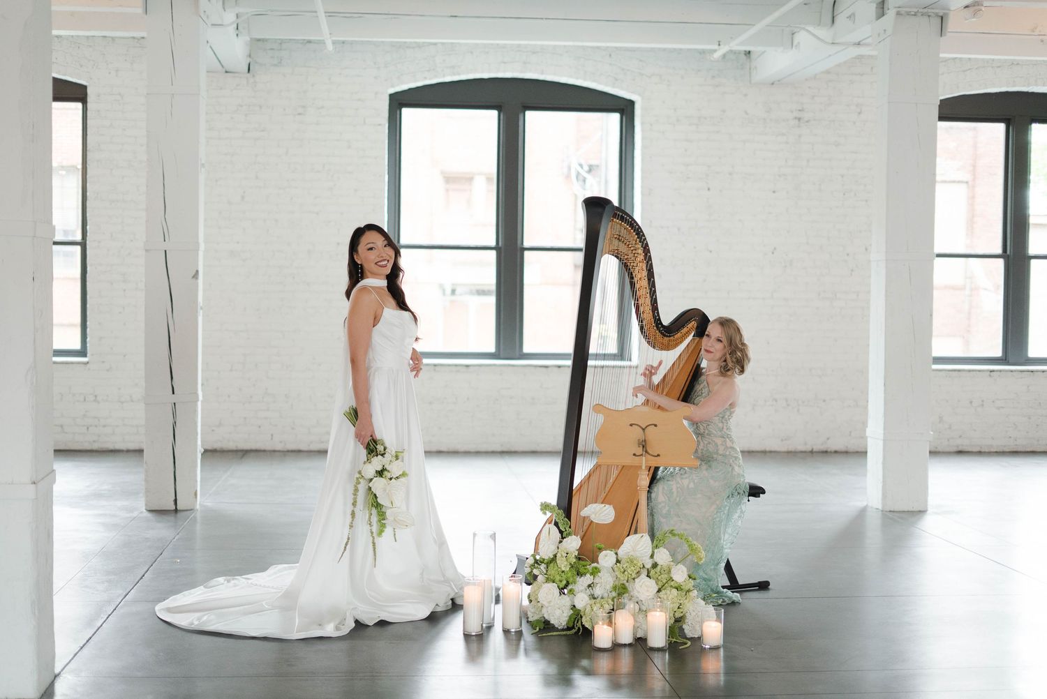 Bride in white gown poses with harpist playing at elegant wedding ceremony in bright white brick venue.