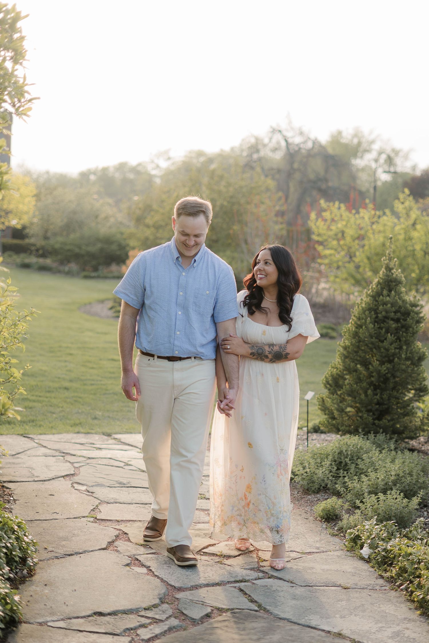 Couple walking hand-in-hand on a stone garden path at golden hour, woman in floral dress, man in blue shirt.