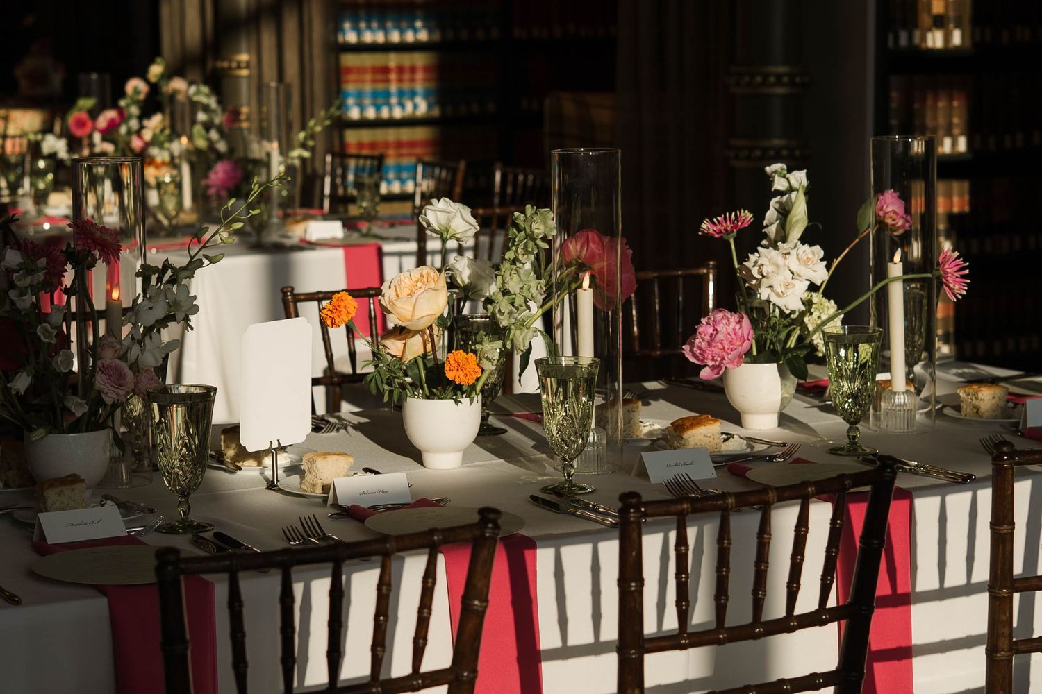 Elegant wedding reception table with colorful floral arrangements, pink ribbon chairs, and candles in a library setting.