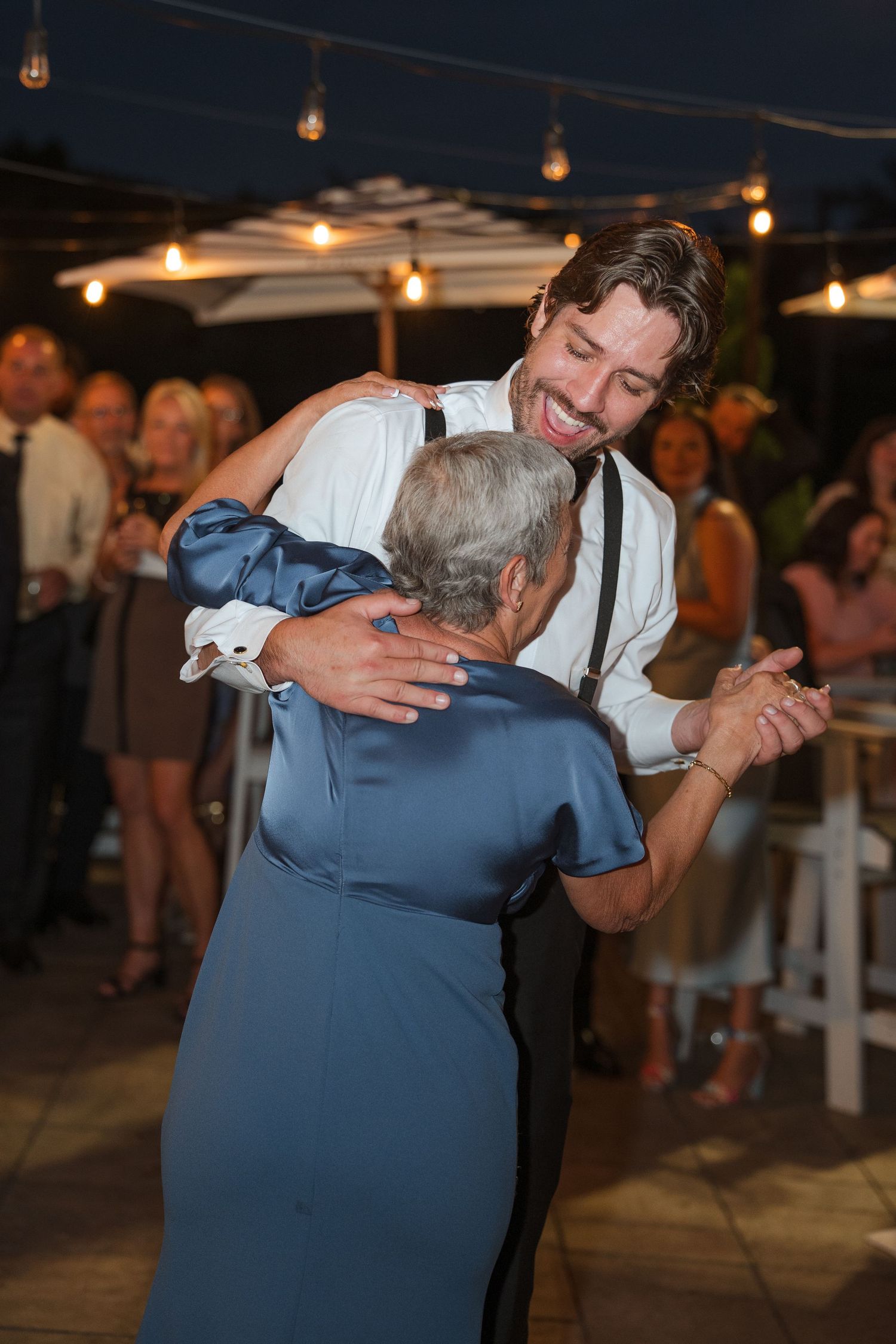 Groom sharing a joyful dance with an elderly guest at an outdoor evening wedding reception under string lights.