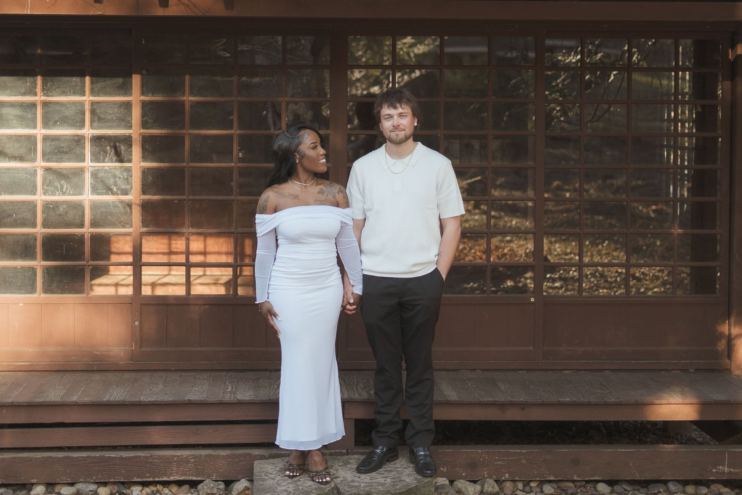 Interracial couple holding hands, posing in front of a wooden building, woman in white off-shoulder gown.