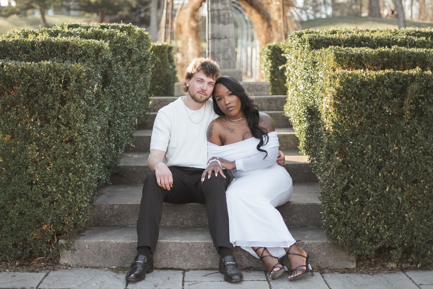 Interracial couple sitting on garden steps, woman in white dress leaning on man in white shirt, surrounded by hedges.