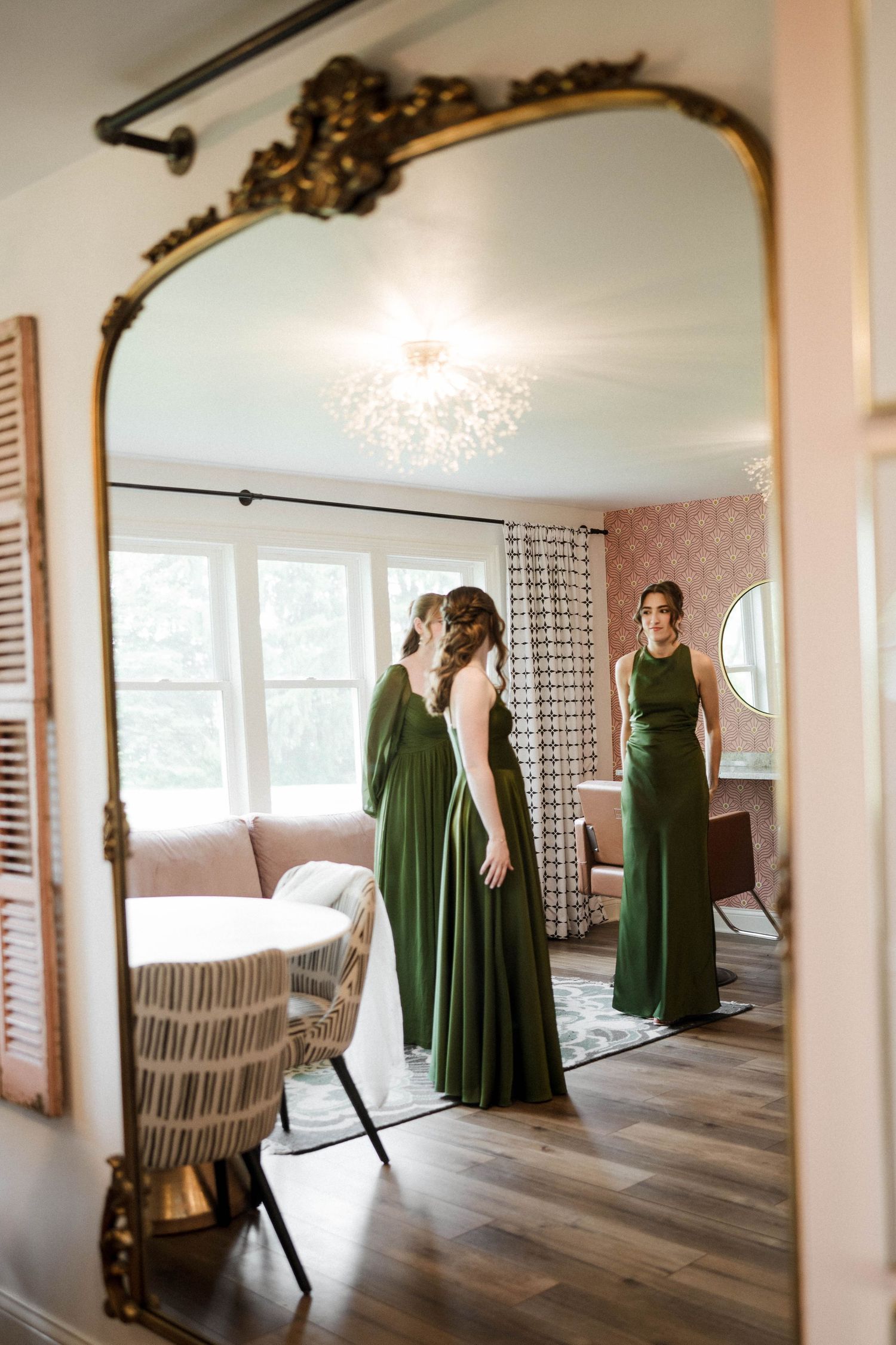 Bridesmaid in elegant green gown admiring her reflection in an ornate gold-framed floor mirror in a pink bridal suite.