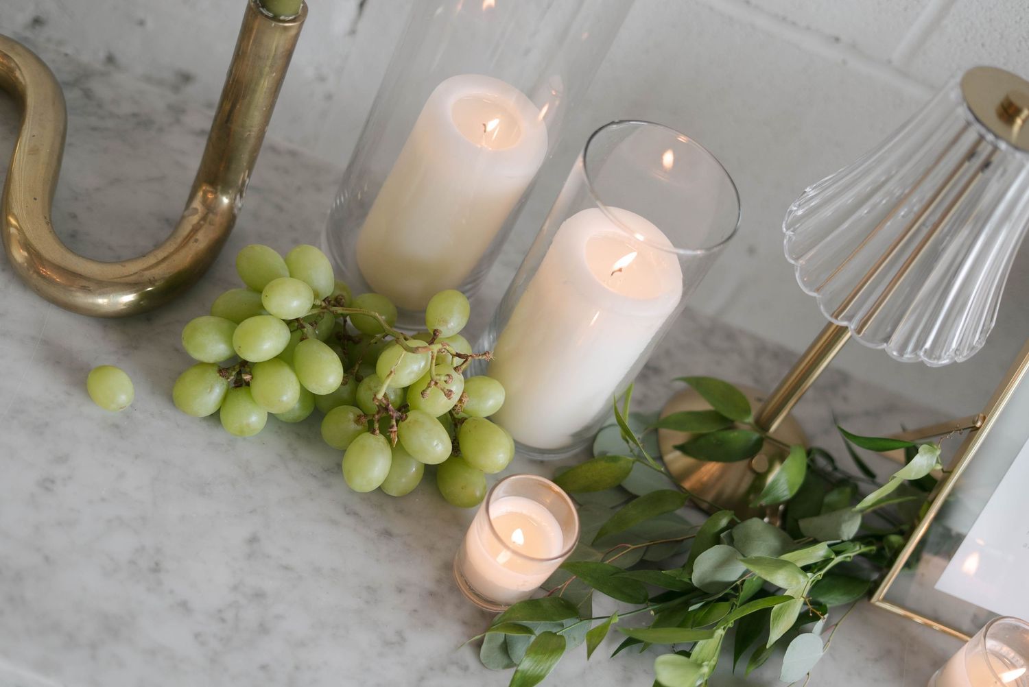 White pillar candles in glass holders with green grapes and eucalyptus on marble surface with gold accents.