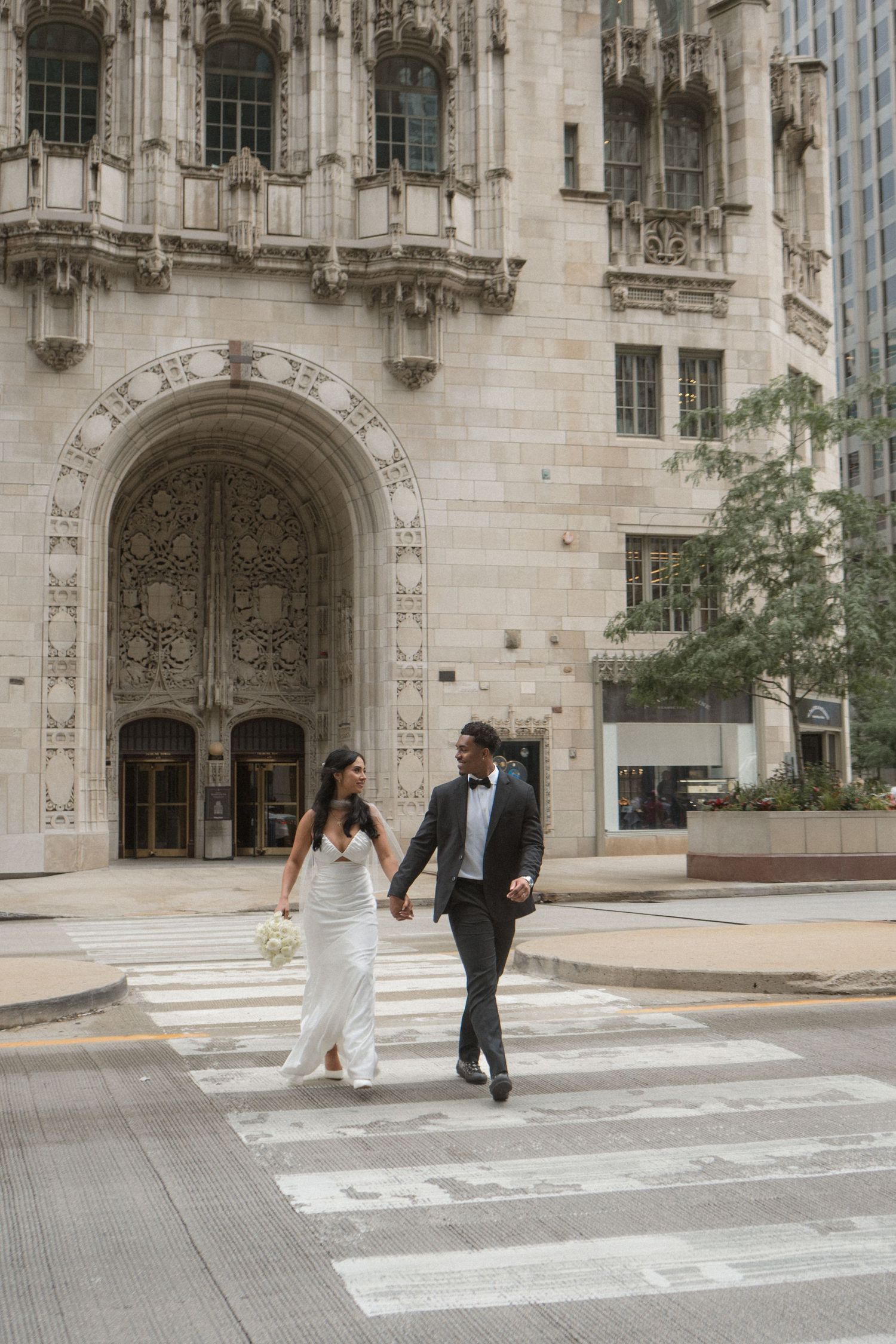 Bride and groom holding hands crossing a zebra crossing in front of an ornate Gothic building in Chicago.