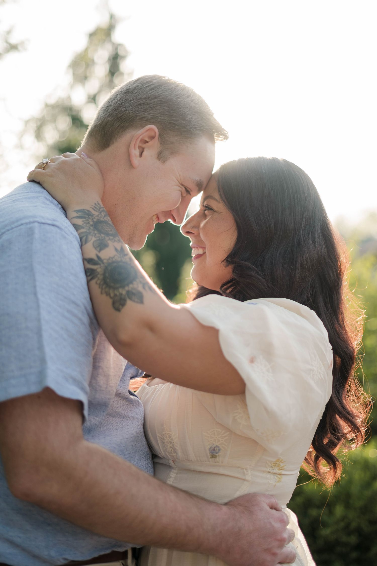 Couple sharing a romantic moment outdoors, man with tattoo embracing smiling woman in white dress, bathed in golden sunlight.