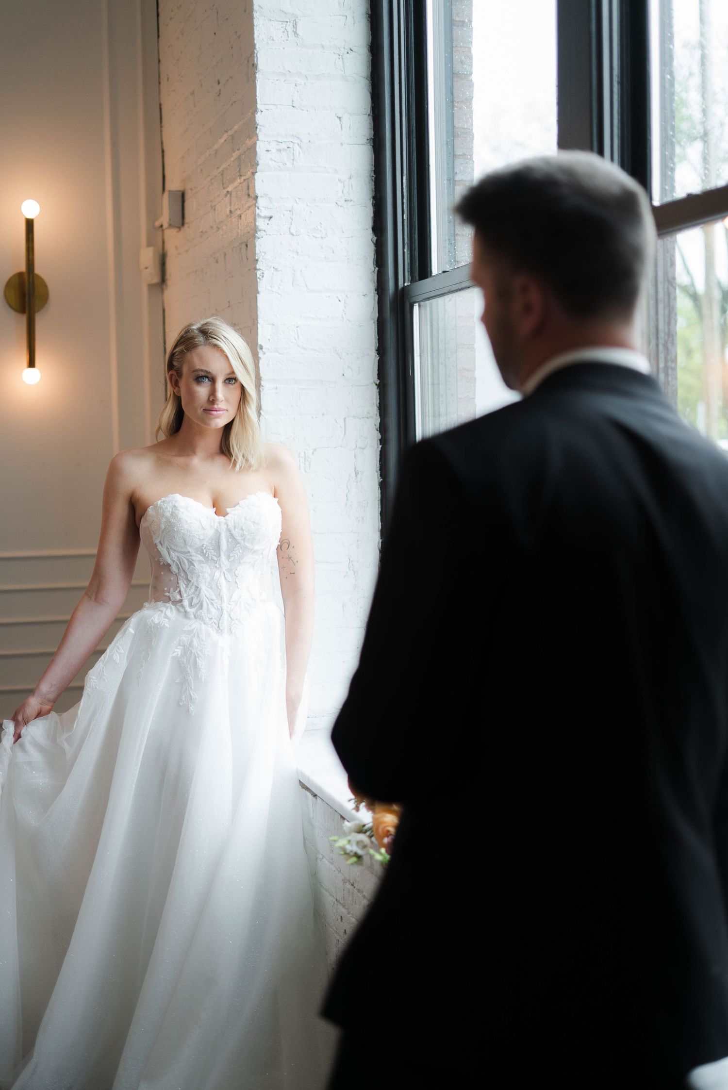 Bride in strapless lace ballgown smiles at groom in black suit during romantic first look by large window.