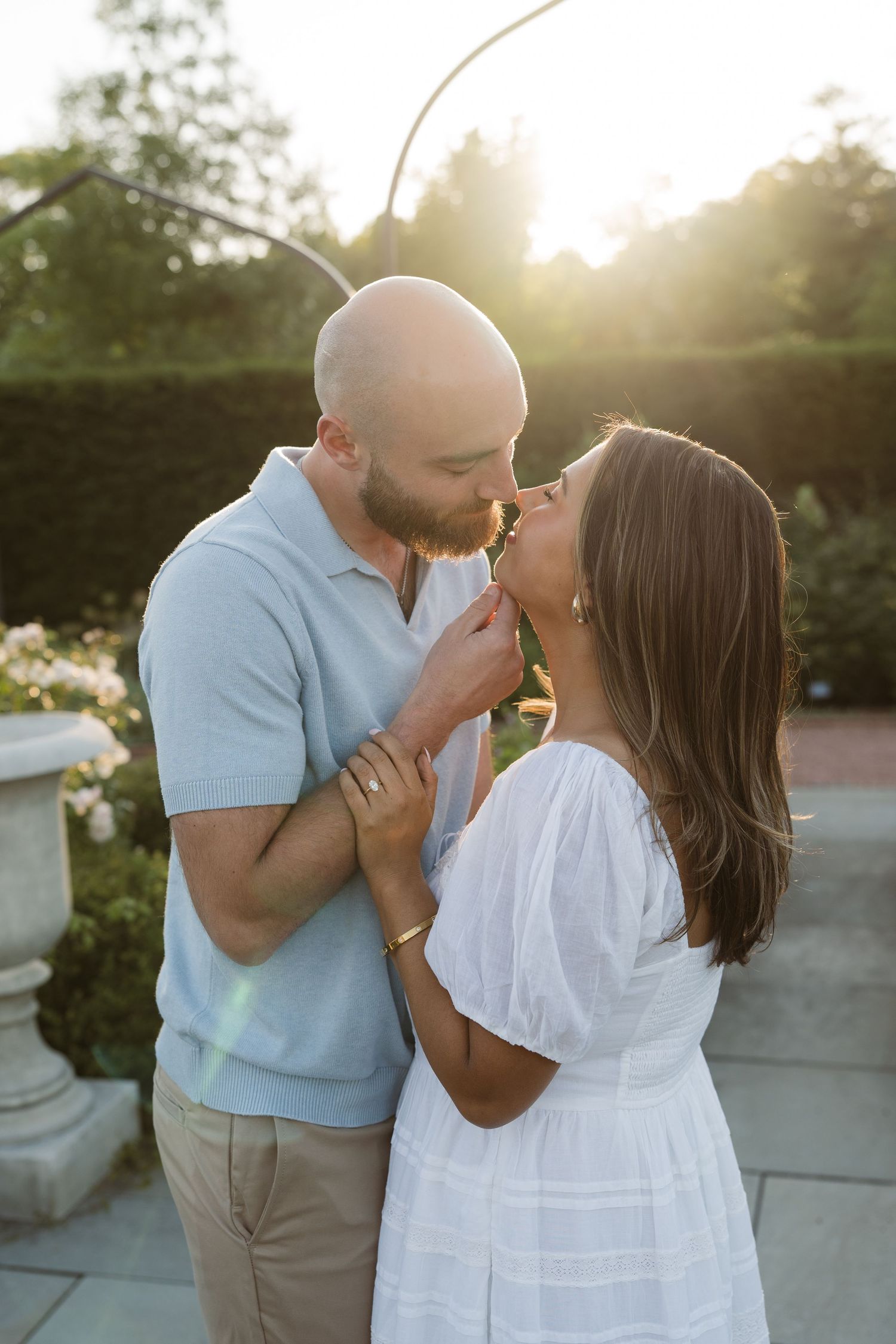 Couple kissing at golden hour sunset in garden, man in light blue polo and woman in white dress holding hands.