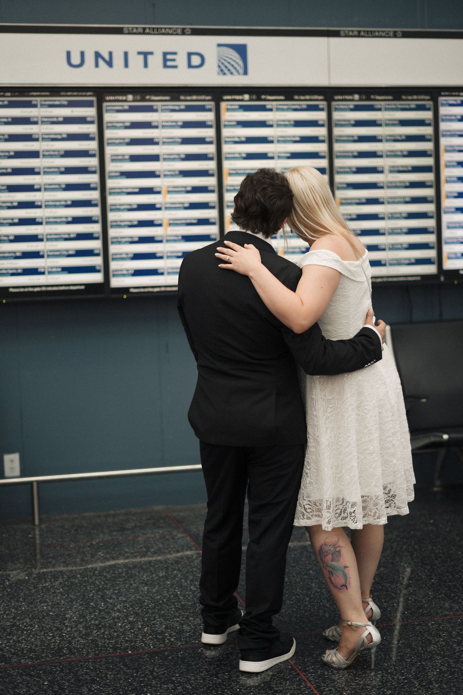 Couple embracing in front of United Airlines flight board at airport, woman in white dress with tattoo, man in black suit.