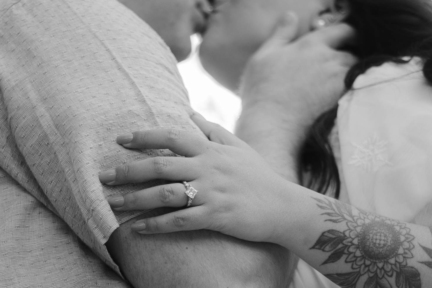 Black and white photo of a couple kissing, woman's hand with engagement ring and floral tattoo resting on man's arm.