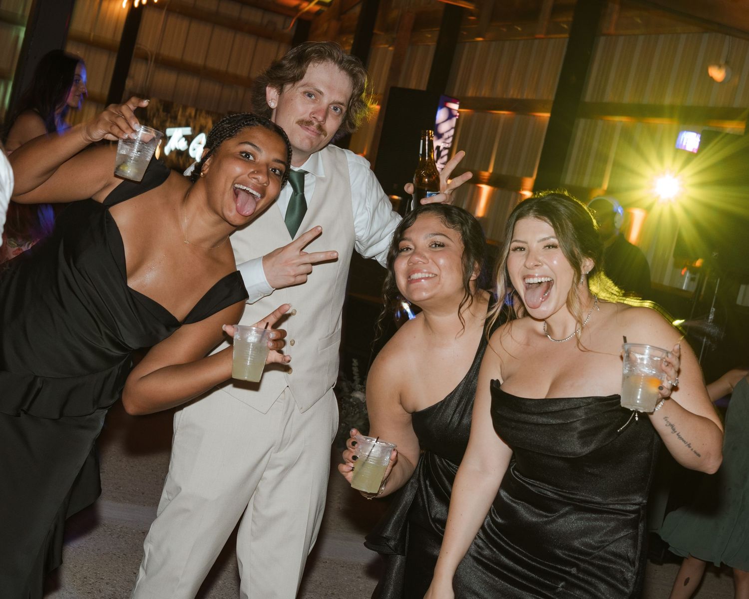 Four friends laughing and dancing together at a formal wedding reception party in a rustic barn venue.