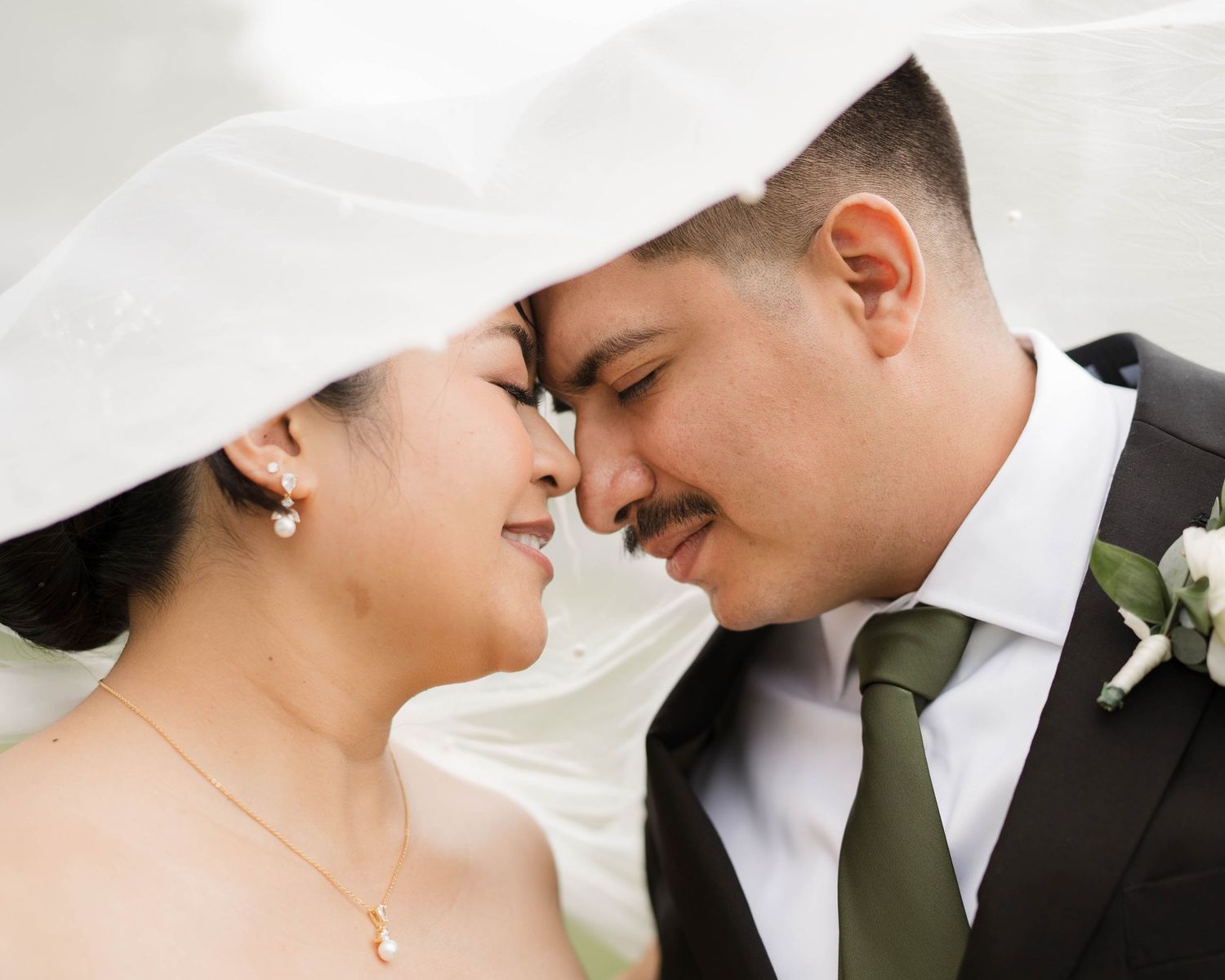 A newlywed couple shares a romantic moment under the bride's veil touching foreheads.