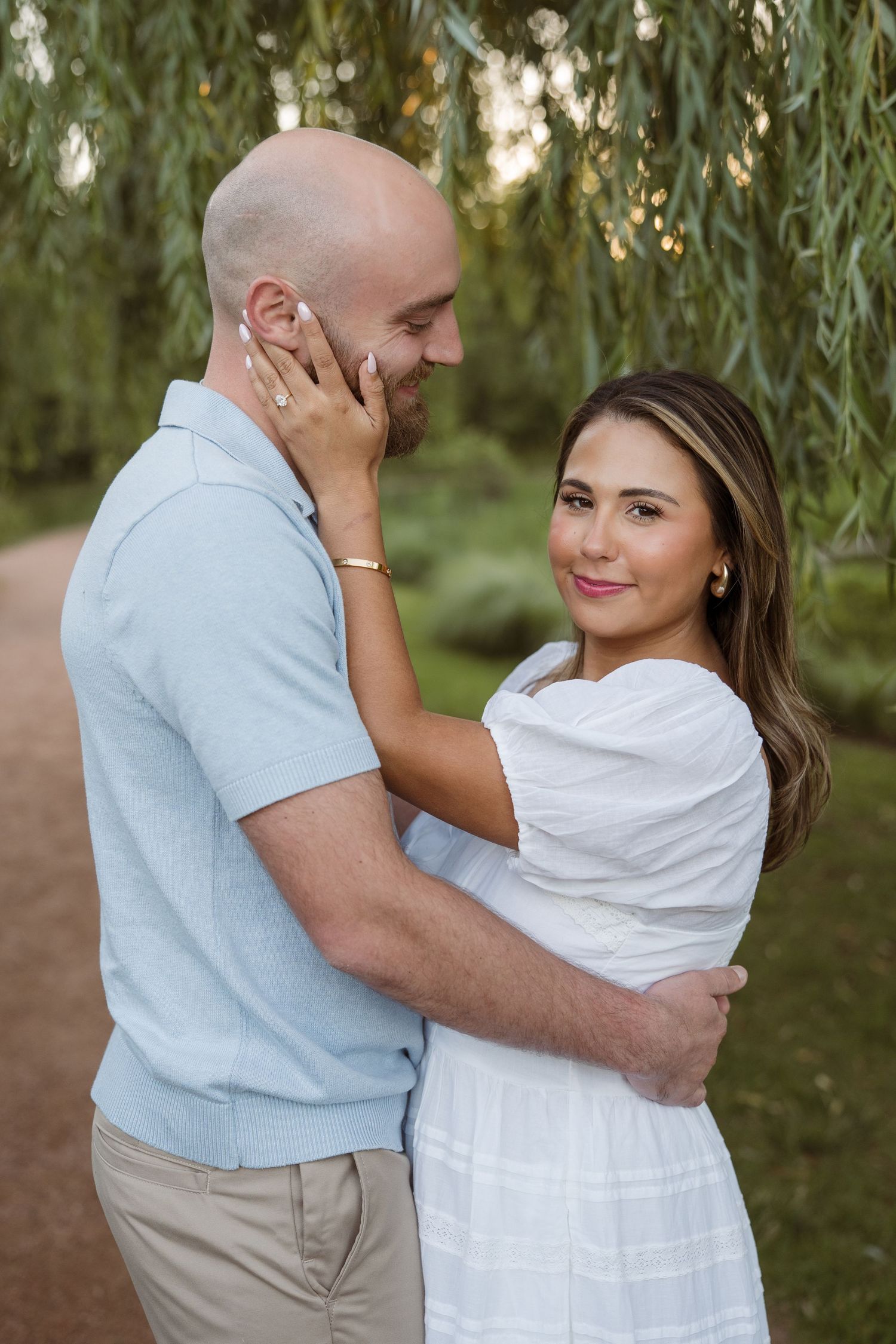 Couple embracing near willow tree, woman in white dress touching mans face during outdoor engagement photo session.